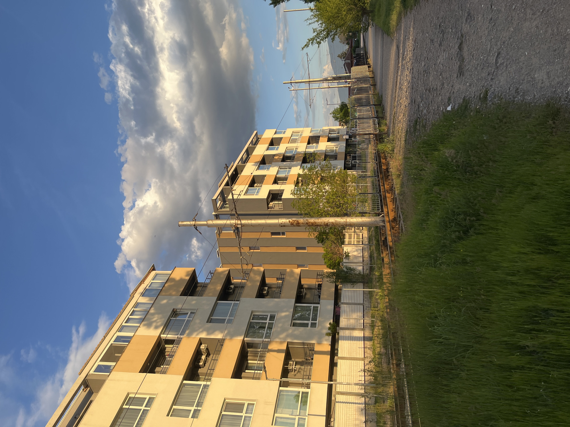 The image shows two modern apartment buildings under a partially cloudy sky, likely located in Sofia, Bulgaria, based on the architectural style. The buildings are multi-story, with alternating beige and grey facades and multiple balconies with white railings. Electrical poles are visible near the buildings and adjacent to a fence line. A dirt and gravel path runs along the edge of the image, bordered by tall green grass. The scene appears to be captured in the late afternoon or early evening due to the warm lighting.