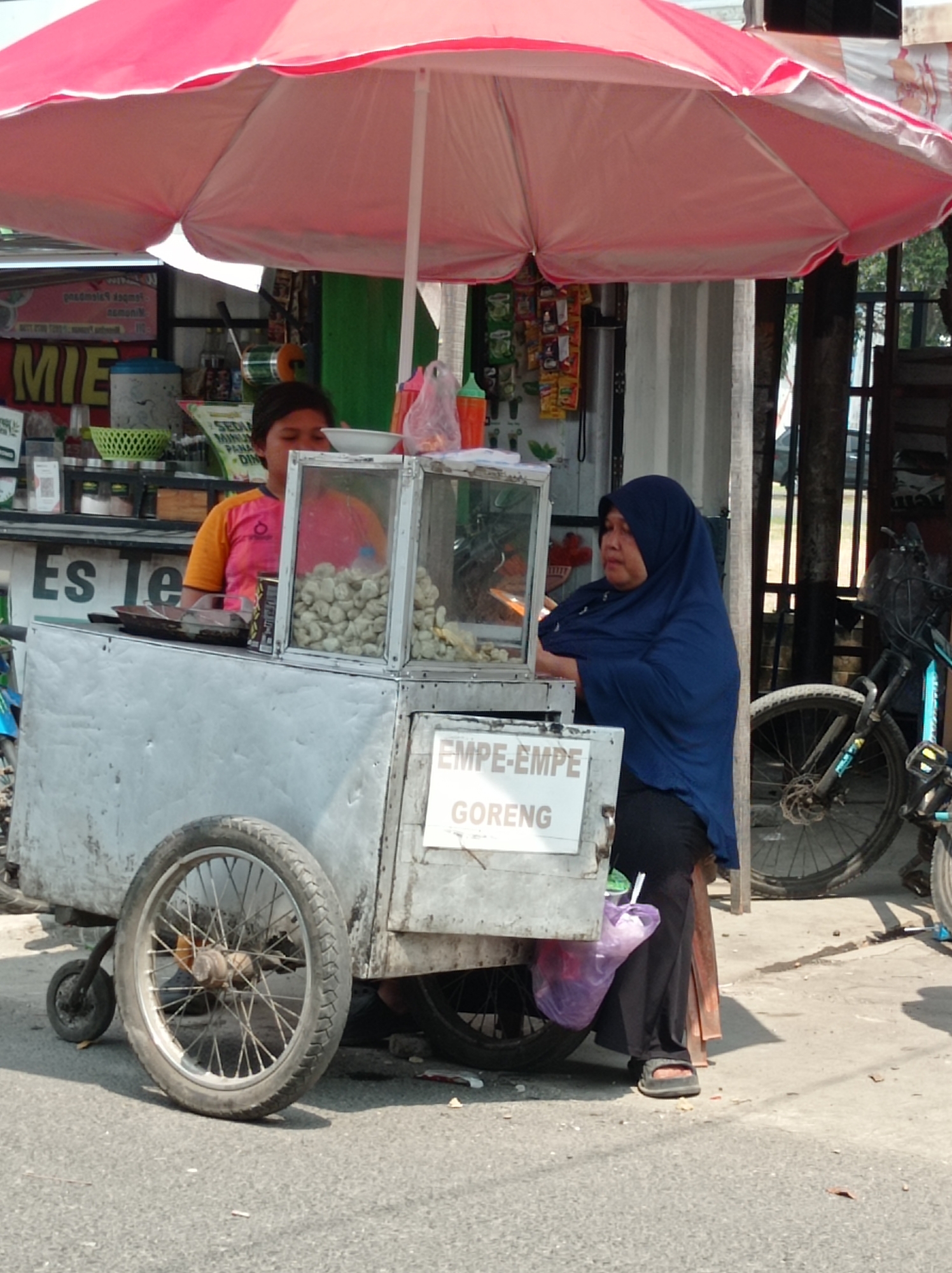 The image depicts a street food cart, possibly in Tegal, Indonesia, a city known for its culinary offerings. The cart is a mobile unit, likely made of metal, and is equipped with a display case for food. A large, pink and white umbrella provides shade over the cart and its occupants.
Two individuals are visible. One, a person wearing an orange and pink t-shirt, stands behind the cart and appears to be attending to the food. They are looking down, possibly at the display case. The second person, wearing a blue hijab and dark clothing, stands to the right of the cart, also seemingly involved in the operation. A bicycle is parked next to the cart, suggesting a mode of transport or perhaps belonging to one of the individuals.
The environment appears to be a street or sidewalk, with asphalt paving visible in the foreground. The background shows elements of a market or commercial area, with stalls and buildings. The overall scene suggests a typical day of street vending, with the focus on the food cart and its operators.