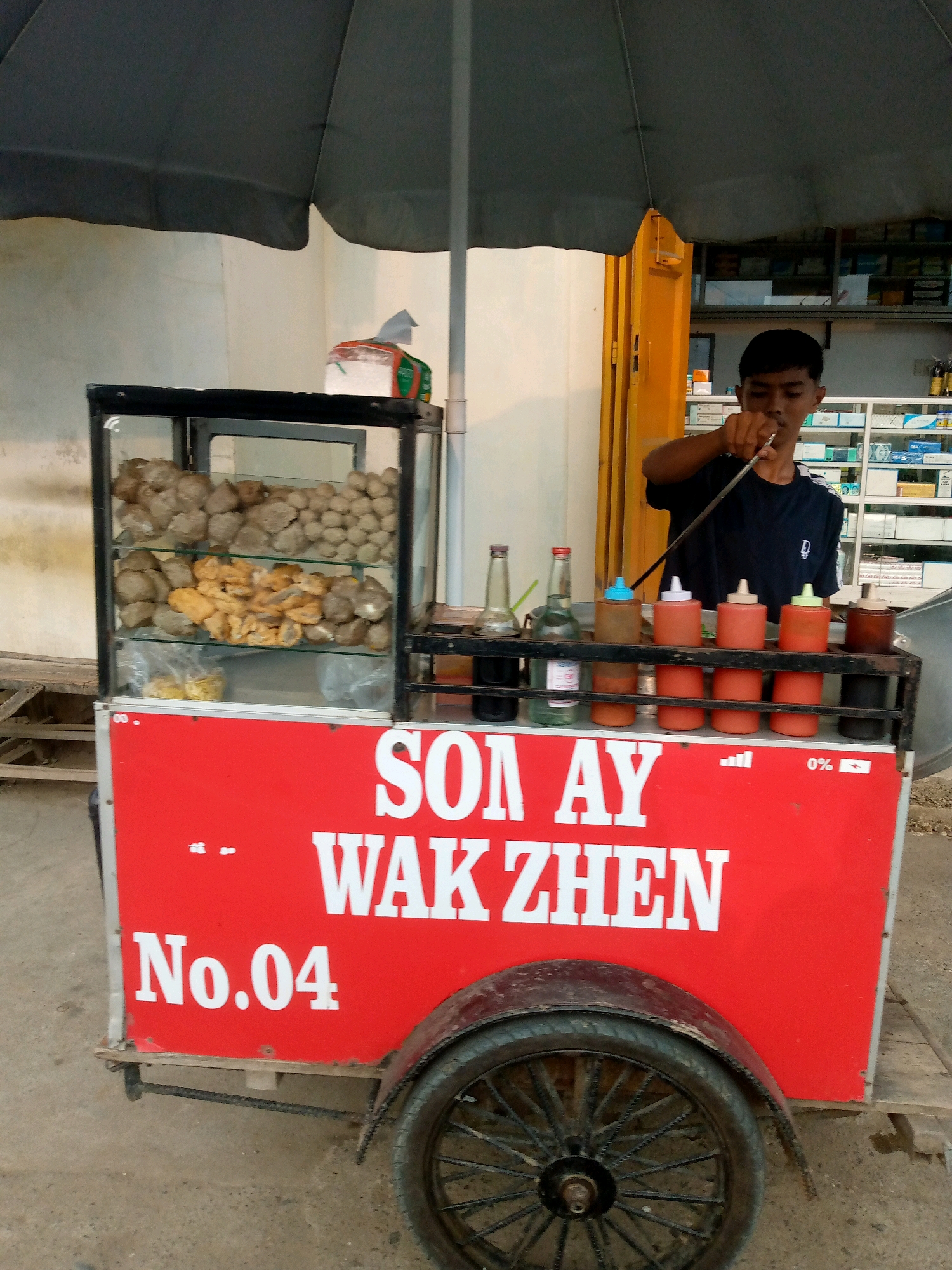 A young man stands behind a food cart with a red exterior and a glass display case. The cart is labeled "SON AY WAK ZHEN No.04". Inside the glass case, there are several trays of food, including what appear to be meatballs and fried items. The man is holding a utensil, possibly for serving food. In front of the cart, there are several bottles and dispensers, likely containing sauces or condiments. The background shows a building with shelves filled with various items, possibly a store or pharmacy. The scene appears to be outdoors, with a large umbrella providing shade over the food cart. The overall setting suggests a street food vendor in Keudegeudong, Indonesia.