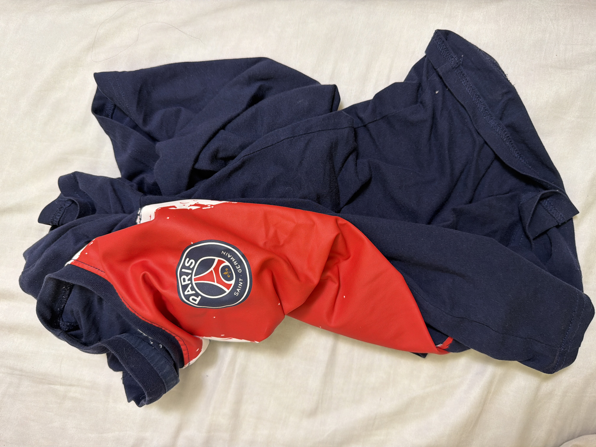 A dark blue t-shirt with a red and white Paris Saint-Germain logo is draped on a white, textured surface, possibly a bedsheet. The t-shirt is bunched and folded, obscuring most of its form. The logo, featuring a circular crest with the Eiffel Tower and the words "PARIS SAINT-GERMAIN," is clearly visible on the red portion of the shirt. The lighting is diffused, casting soft shadows. The image is taken from a top-down perspective.