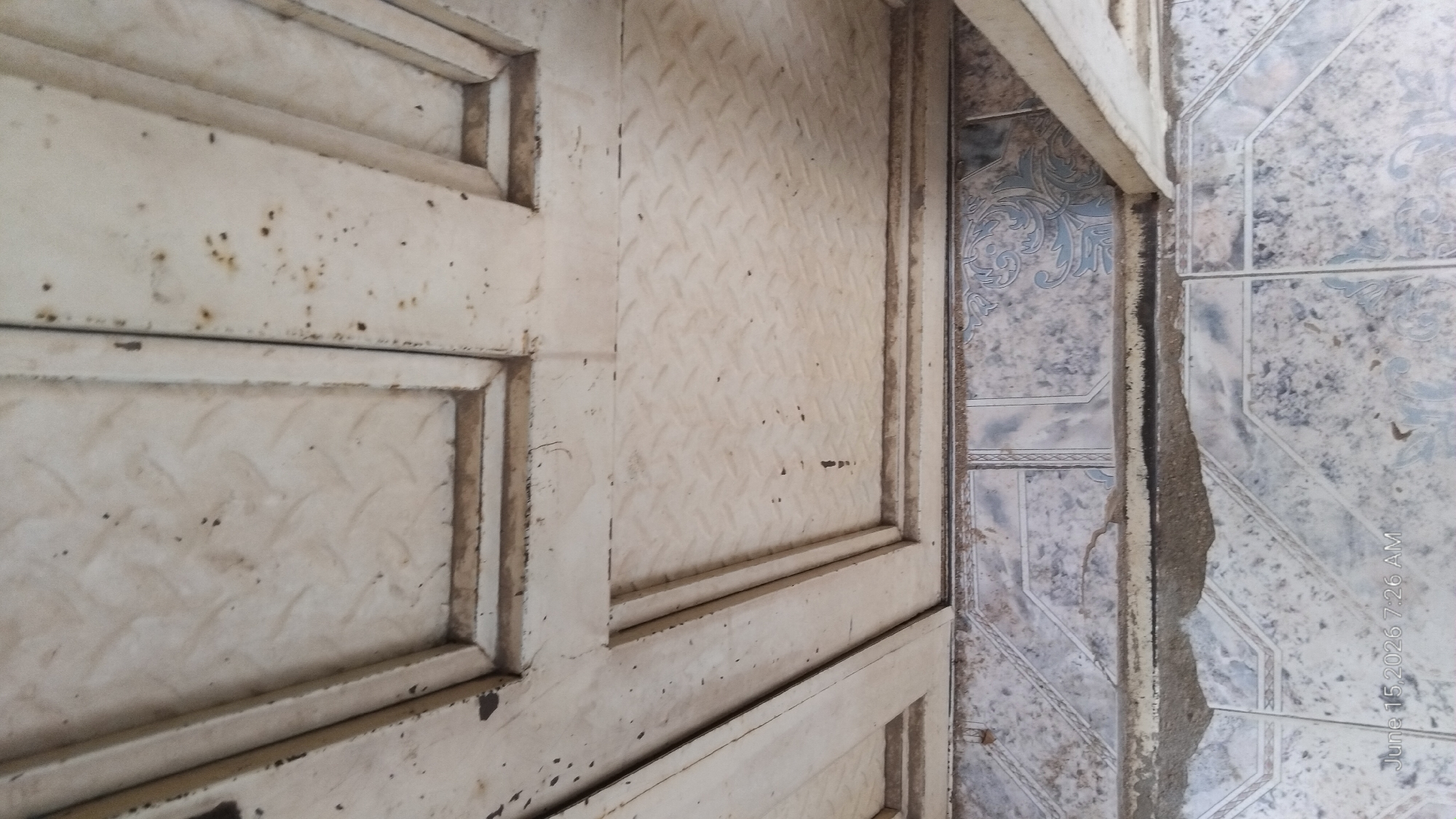 The image depicts a close-up, low-angle view of the lower section of what appears to be an old, weathered metal door or gate and the tiled floor beneath it. The metal surface, painted a faded off-white, is textured with a diamond plate pattern and shows significant signs of age, including rust spots and peeling paint. The door is divided into panels by raised metal strips. Below the door, the floor is covered in square tiles with a patterned design in shades of blue and grey, set against a speckled white background. Some of the tiles appear cracked or worn. The timestamp "June 15, 2026 7:26 AM" is visible in the lower left corner, suggesting the image was taken on this date and time. The scene conveys a sense of disrepair and urban decay. The location in the city of Howayi, Nigeria, implies a specific context for this observation.