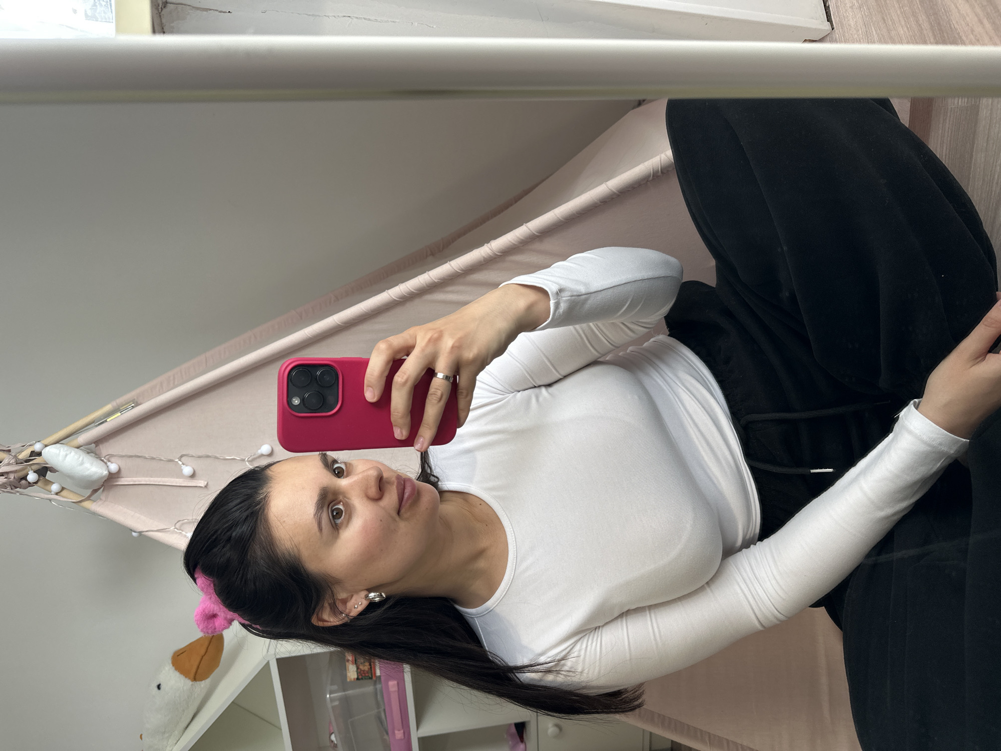 The image is a mirror selfie featuring a woman indoors. She is positioned on her side within the frame, reflecting the image's original orientation. The woman has long dark hair, dark eyes, and is wearing a white long-sleeved top and black bottoms. A pink fluffy hair accessory is visible in her hair. She holds a smartphone with a bright pink case and a triple-camera setup in her right hand, aimed at her reflection. A silver ring is worn on her right ring finger.
The setting appears to be a cozy indoor space, possibly a child's room or a play area. A pink tent-like structure, possibly a teepee, adorned with small string lights, is prominent in the background. A white plush toy, resembling a goose with an orange beak, is partially visible near the bottom left corner. A white shelf with clear storage containers is also visible behind her. The lighting is bright and even, characteristic of an indoor environment. The woman has a neutral expression, looking directly towards the phone's camera. There is no visible text in the image.