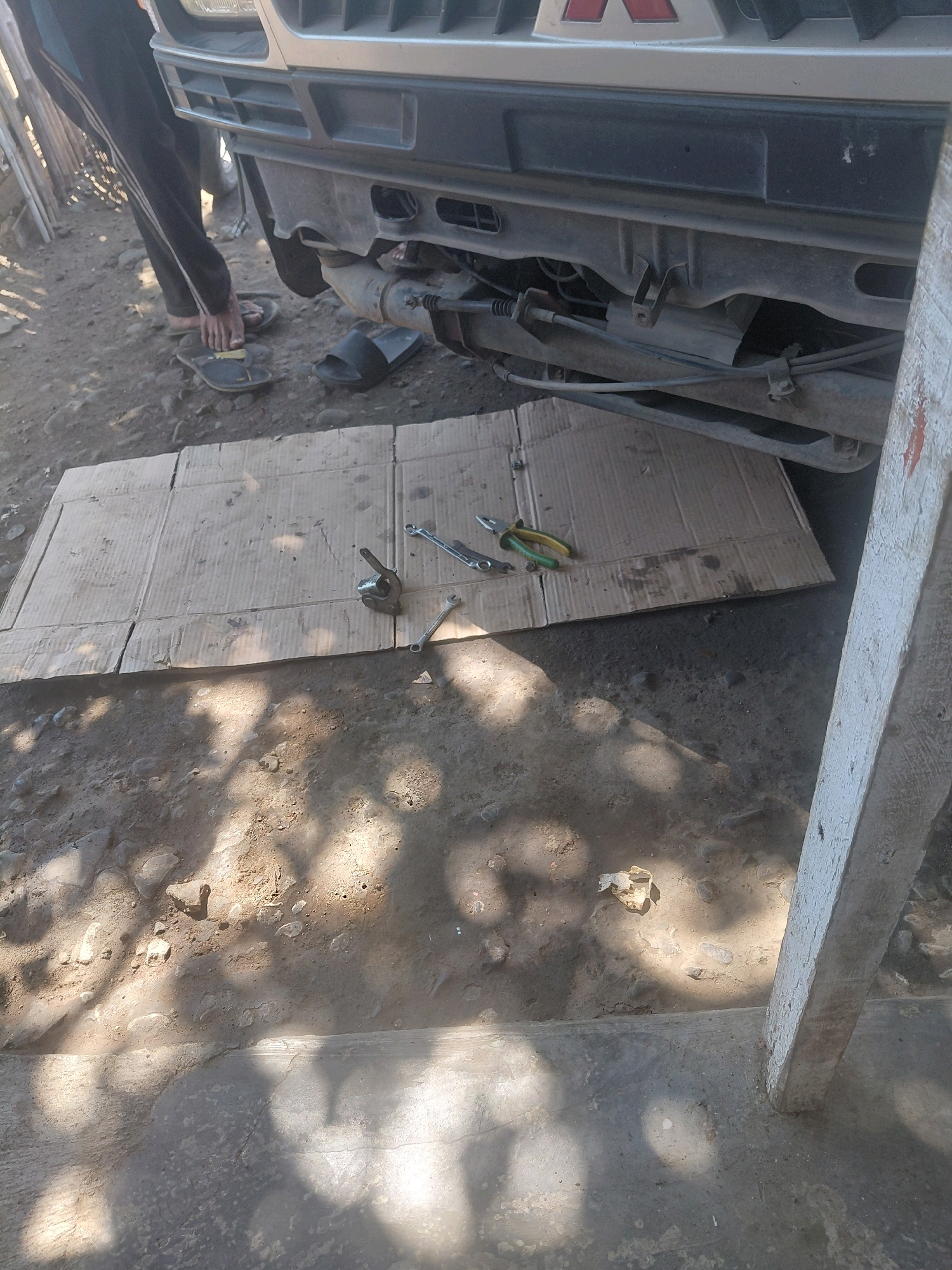 In Paiton, Indonesia, a person wearing dark track pants and flip-flops is seen working on the underside of a vehicle. The vehicle appears to be a light-colored SUV or similar model, with its front bumper and chassis exposed. A piece of flattened cardboard is laid out on the ground, serving as a makeshift work surface. On the cardboard, various tools are placed, including a pair of pliers with green and yellow handles and at least two wrenches. The ground is a mixture of dirt, gravel, and some concrete. The scene is outdoors, with shadows cast by trees or other overhead structures suggesting it is daytime. Another pair of black flip-flops lies near the car's undercarriage.