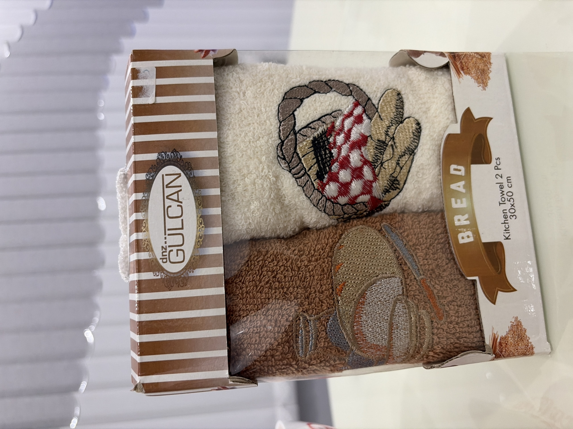 The image shows a product display of two kitchen towels packaged in a clear plastic box with a decorative cardboard sleeve. The sleeve is brown and white striped with a label that reads "dnz... GULCAN". The towels inside have embroidered designs. The left towel is brown and features an embroidered image of a loaf of bread next to a knife. The right towel is cream-colored and has an embroidered image of a basket with a red and white checkered cloth inside. Below the towels, a brown ribbon banner reads "BREAD" and text beneath it indicates "Kitchen Towel 2 Pcs 30x50 cm". The product is set against a blurry white background, possibly a curtain or wall. There are no people visible in the image. The location in the city of Podgumer, Bulgaria is not discernible from the visual information provided.