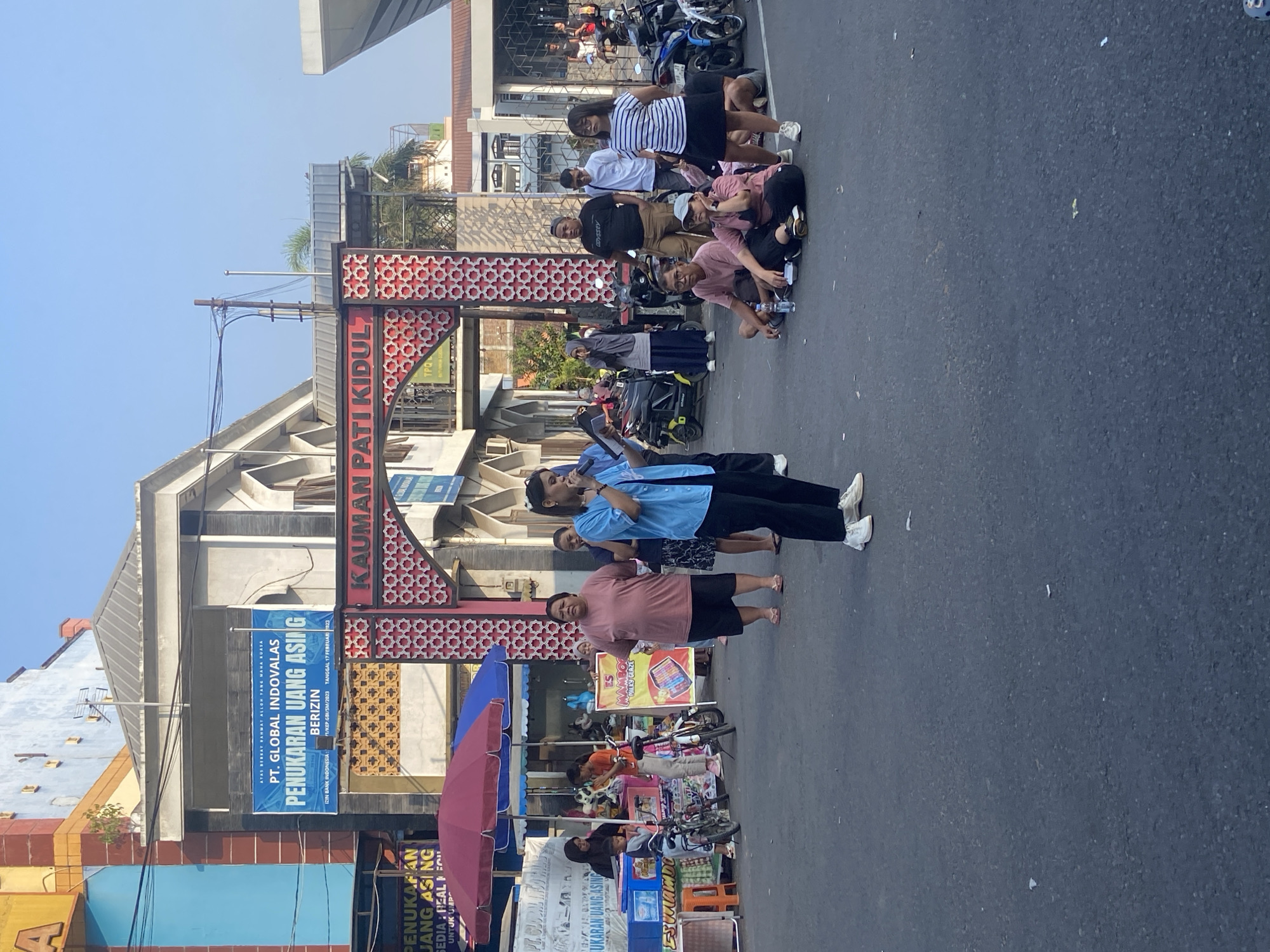 In a street scene in the city of Kecamatan Pati, Indonesia, a group of people are gathered. In the foreground, a woman wearing a blue shirt and black pants is speaking into a microphone. Behind her, other individuals are seated on the ground, some appearing to listen. To the left, a market stall with colorful awnings is set up, with people attending to it. Further back, a large arched gateway with intricate red and white patterns is visible, likely an entrance to a building or area. Various motorcycles are parked in the background. The overall environment suggests an outdoor public gathering or event on a bright, clear day.