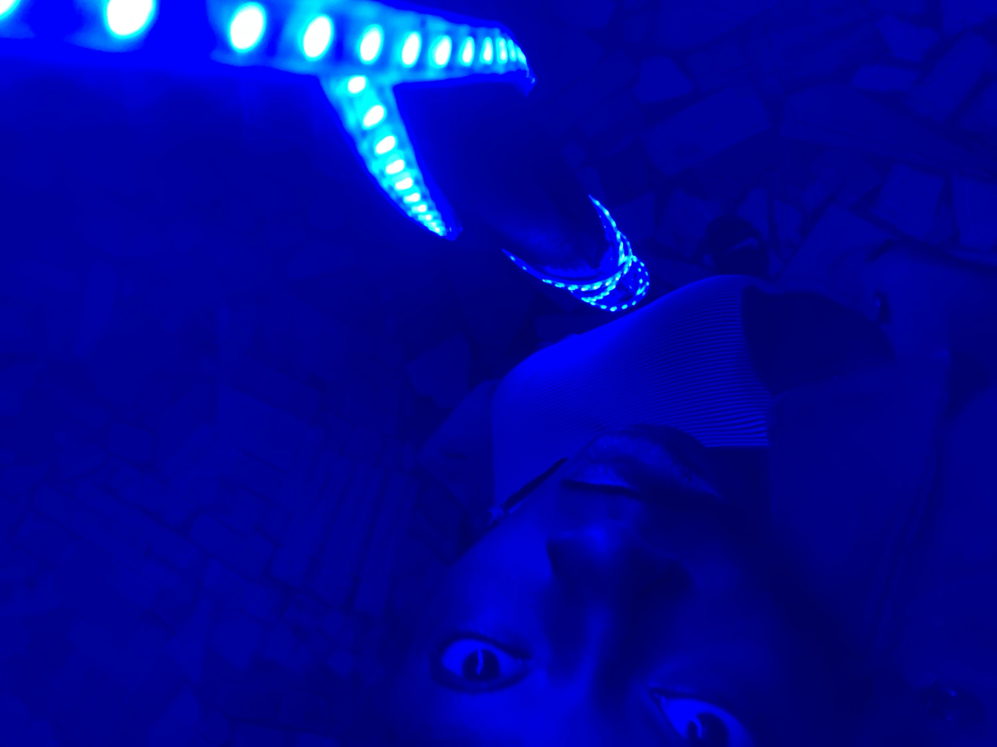This low-light photograph captures a scene bathed entirely in vibrant blue light, suggesting a nighttime or indoor setting. The primary subject is a person, whose face, including prominent eyes and parts of their nose and mouth, is visible in the lower center of the frame. They appear to be looking directly at the viewer, possibly from a reclined or upward-facing position. The person seems to be wearing a light-colored, possibly striped, top.
Dominating the upper part of the image are bright, cool blue LED strip lights. One strip forms a distinct 'T' or cross shape, originating from the top left and extending across the frame. Another segment of similar blue LED lights is wrapped around a cylindrical object, possibly an arm or another fixture, on the right side.
The background behind the person and to the right features a textured surface, resembling a wall or ceiling adorned with an irregular pattern of rectangular or block-like elements, possibly tiles or decorative panels, all heavily tinted blue by the ambient lighting. The strong blue illumination creates a mysterious, futuristic, or immersive atmosphere. No specific activity beyond posing for a picture is clearly discernible. The scene appears to be set in an indoor environment, potentially within a venue like a club, lounge, or themed room, consistent with a general location context of 'New Stadium, Nigeria,' though no direct stadium features are visible. No visible text is present in the image.
