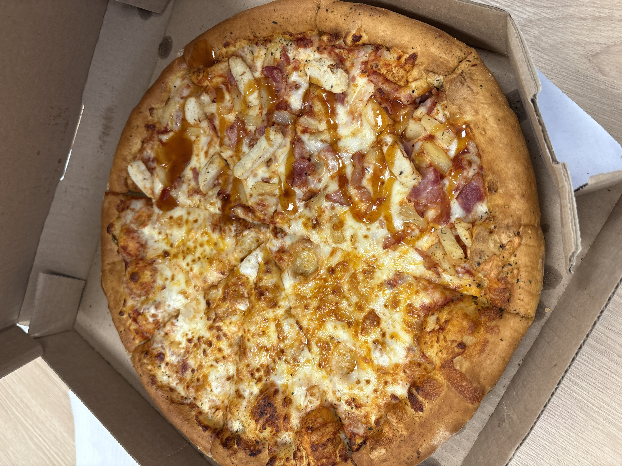 An overhead shot captures a large, freshly baked pizza presented in an open, brown cardboard pizza box. The pizza, occupying the majority of the box, showcases a distinct split of toppings, dividing it into two halves.
The upper half of the pizza features a more elaborate combination of toppings, including generous chunks of white meat chicken, golden pieces of pineapple, and bits of what appears to be bacon or ham, all generously drizzled with a glossy, reddish-brown sauce that suggests a sweet and tangy flavor, possibly barbecue. Melted white cheese and some herbs are visible beneath and among these toppings.
In contrast, the lower half of the pizza is a classic cheese variant, covered with ample melted cheese that has developed appealing browned, bubbly spots, indicating a well-cooked finish. The crust encircling the entire pizza is thick, golden-brown, and appears to be hand-tossed or stuffed, with specks of dried herbs visible on its surface.
The pizza box itself is a standard delivery box, showing some light grease stains on the cardboard interior. The box rests on a light-colored wooden surface, possibly a table or countertop, with the subtle grain of the wood visible beneath. No people are visible, and no text is discernible in the image. The scene suggests a meal ready to be enjoyed in Mexico City, Mexico.