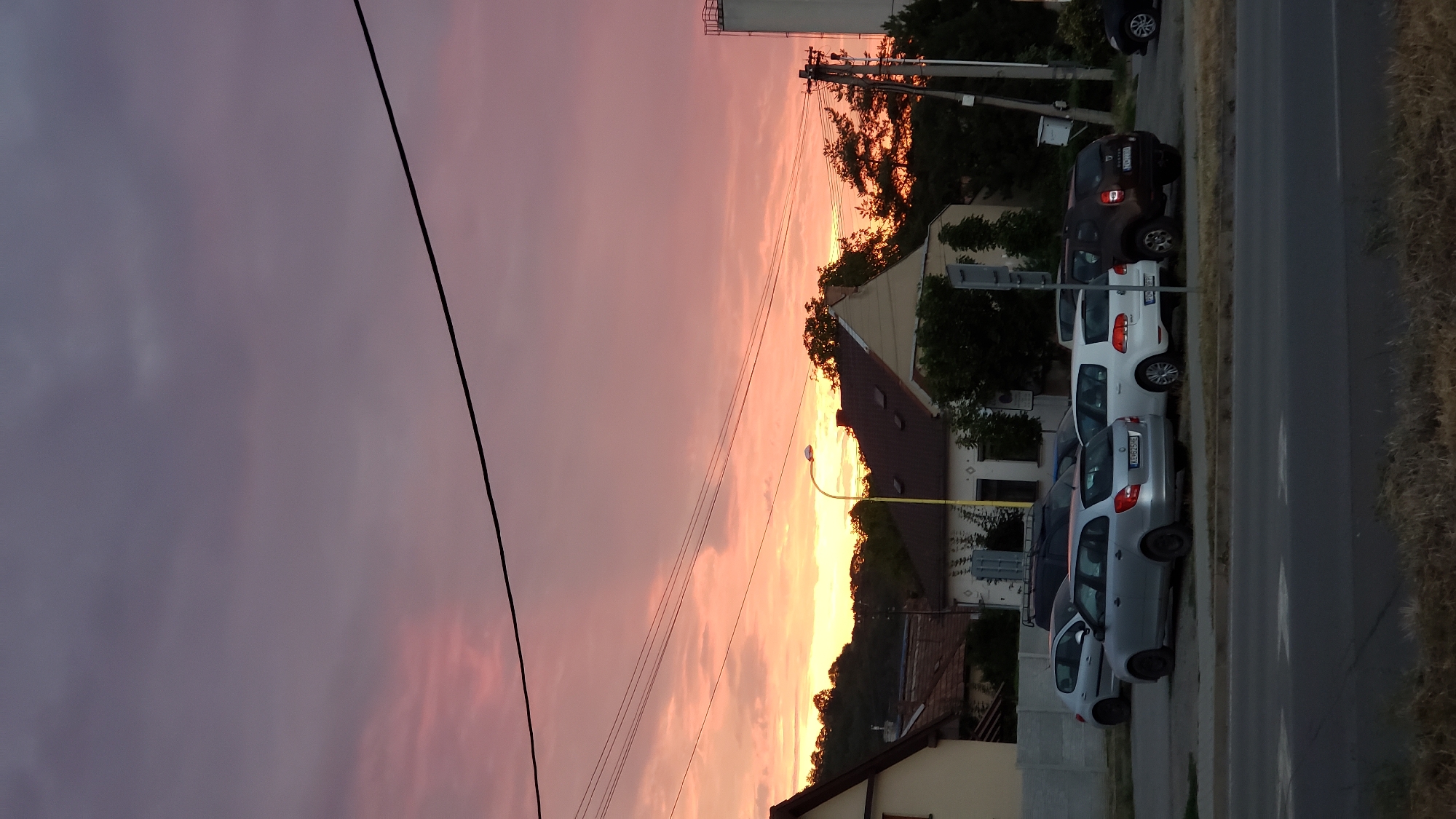 The image depicts a twilight scene in the city of Prešov, Slovakia. The sky is filled with a dramatic sunset, showcasing vibrant hues of pink, orange, and purple, with clouds diffused across the expanse. In the foreground, a street is visible with dry grass on its side. Beyond the street, a row of parked cars is situated in front of a residential area. The cars include a silver station wagon, a white hatchback, and a dark brown SUV. Several houses with pitched roofs are visible behind the cars, interspersed with trees. Utility poles with wires stretch across the scene, one with a street lamp attached. In the background, a gentle hill is covered with dense foliage. The overall atmosphere is calm and serene, capturing the transition from day to night in an urban setting.