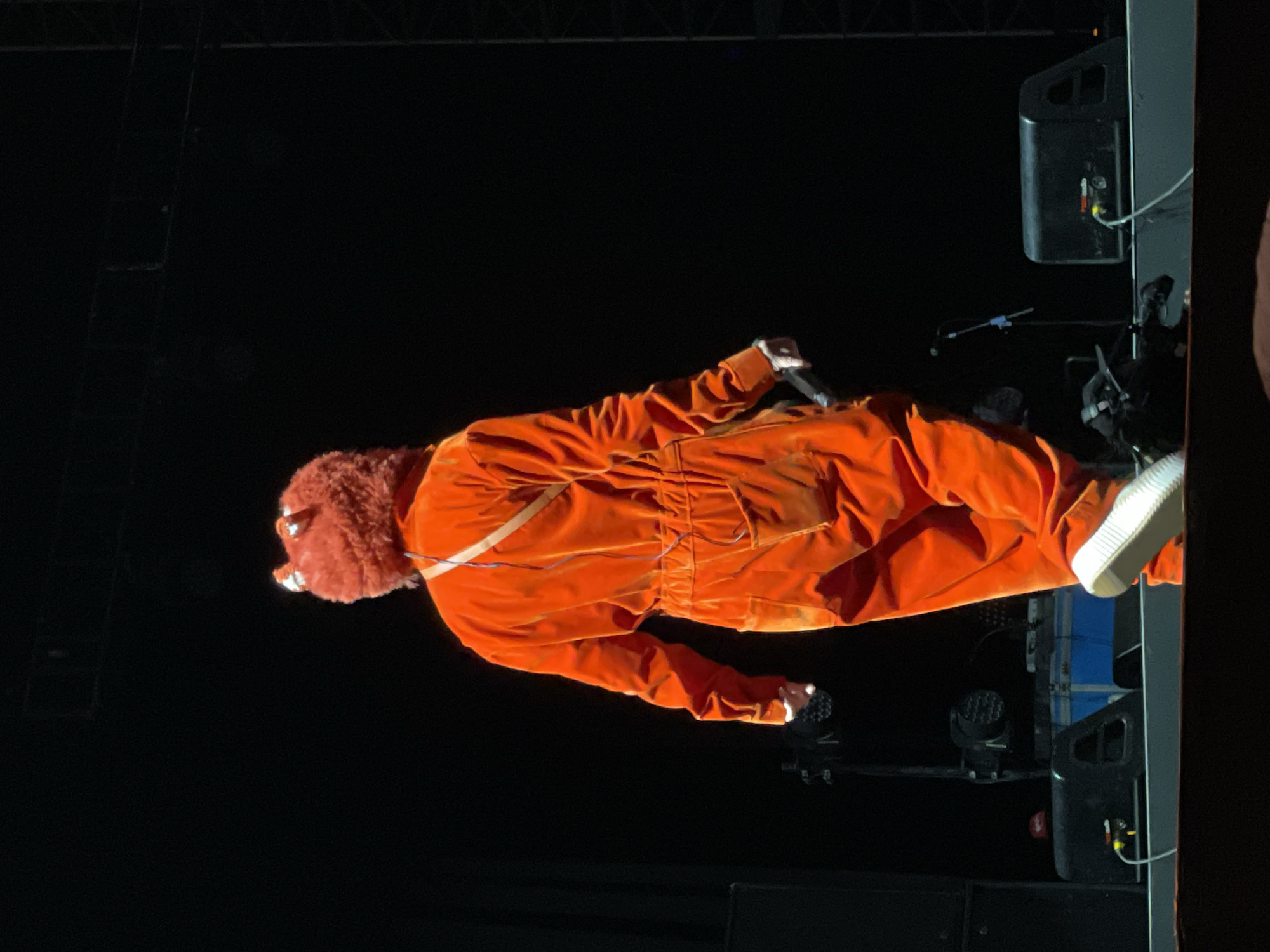 On a dark stage in Salembaran, Indonesia, an individual wearing an orange jumpsuit and a furry brown bear head stands near a large black speaker. The person is holding a microphone. A lighting rig is visible in the background.