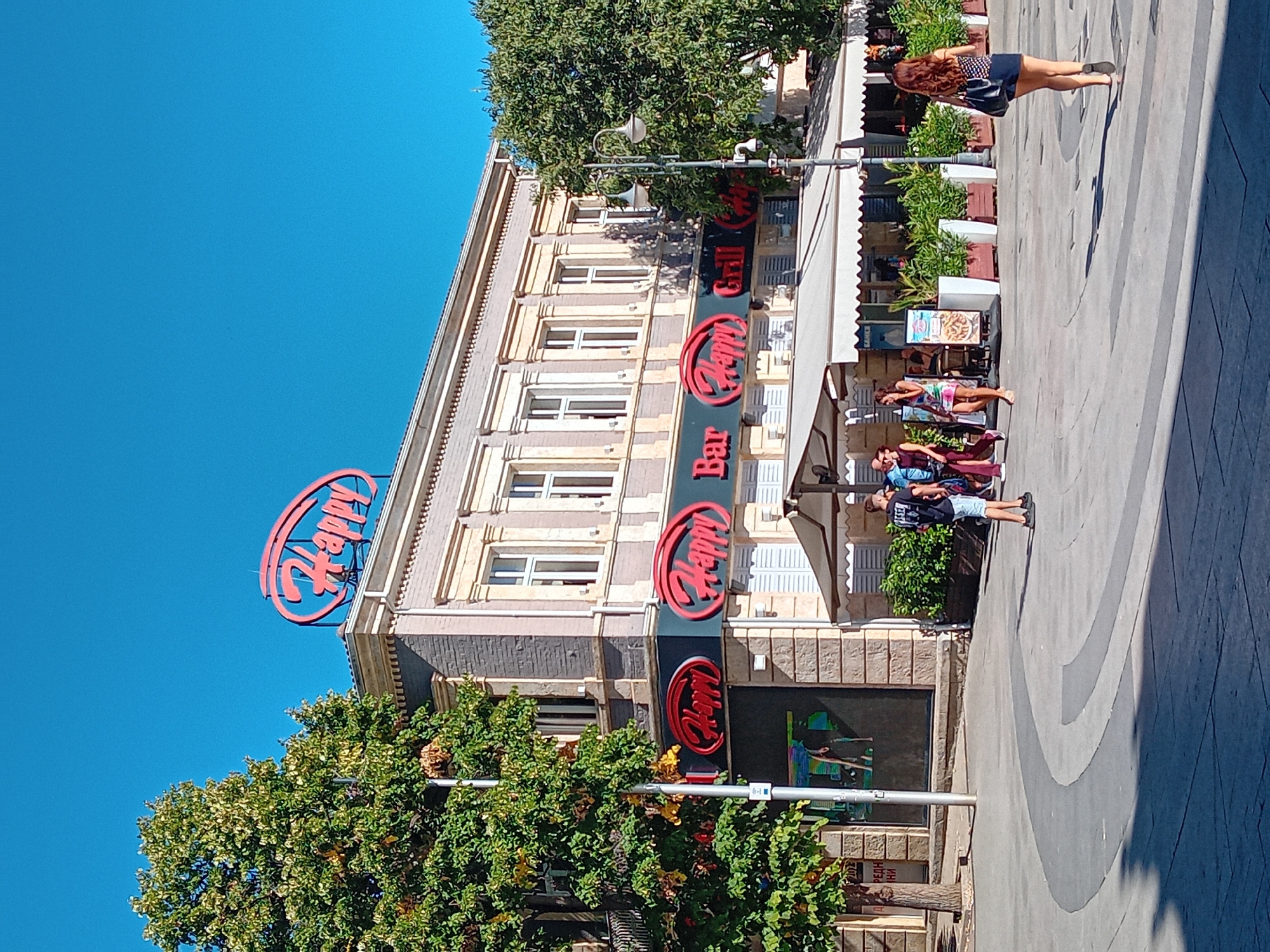 The image shows the Happy Bar & Grill restaurant located in the city of Varna, Bulgaria. The building is a multi-story structure with a distinctive red oval "Happy" sign on its rooftop and another "Happy Bar Grill" sign on its facade. The facade is adorned with classical architectural elements such as pilasters and decorative cornices. In front of the restaurant, there are several white awnings, outdoor seating areas with plants in planters, and a street lamp. Several people are visible in the scene: a woman walking away from the camera towards the right, a group of people walking towards the left, and a man standing. The scene is set on a paved pedestrian area with a curved design. The sky is clear blue.