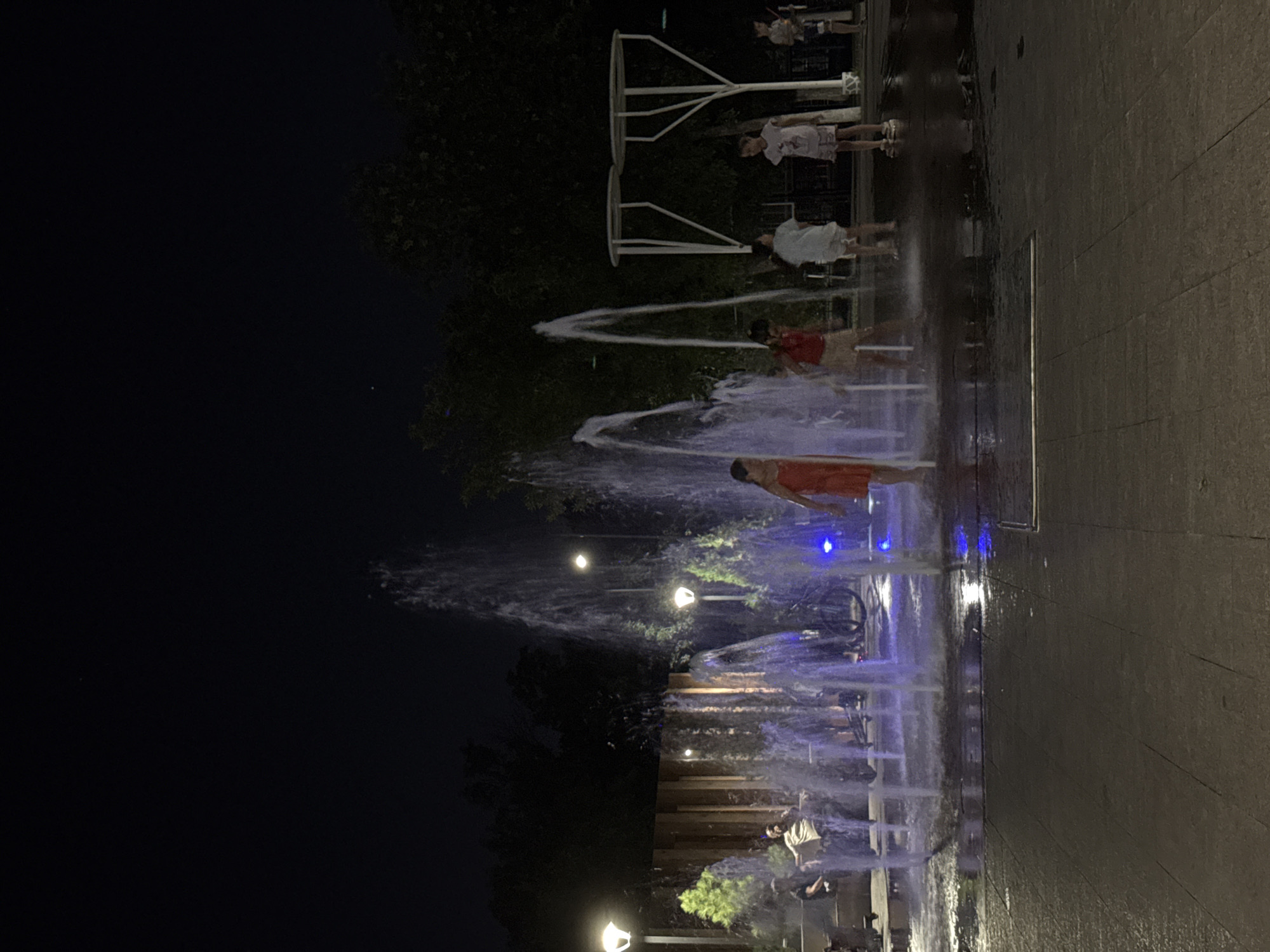 This is a low-light, nighttime photograph of a water fountain in a public park, likely in Shymkent, Kazakhstan. Several children are playing in the fountain, with water spraying around them. The fountain jets are illuminated with blue lights. In the background, trees and park lighting are visible, along with some people sitting on benches. The ground appears to be paved, and it is wet from the fountain's spray. A bicycle is also visible near the fountain. In the distance, there are some modern white structures that appear to be part of the park's architecture.