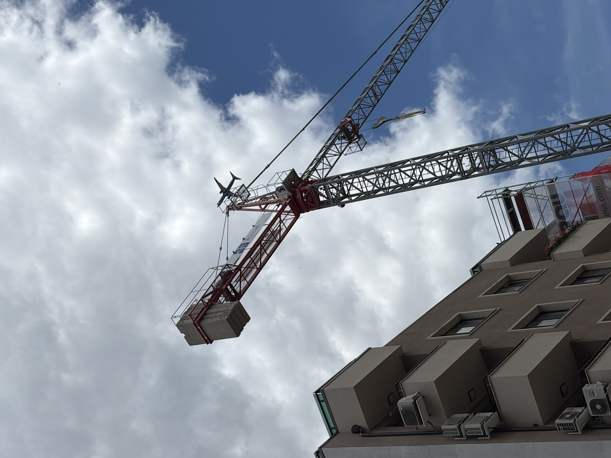 The image shows a construction site with a tall crane and a multi-story building, under a cloudy sky. The building is light brown with balconies and air conditioning units. The crane is red and grey, with its boom extended, and appears to have a model airplane attached to its jib. A banner on scaffolding reads "ROU unit.com". The scene is located in Sofia, Bulgaria.