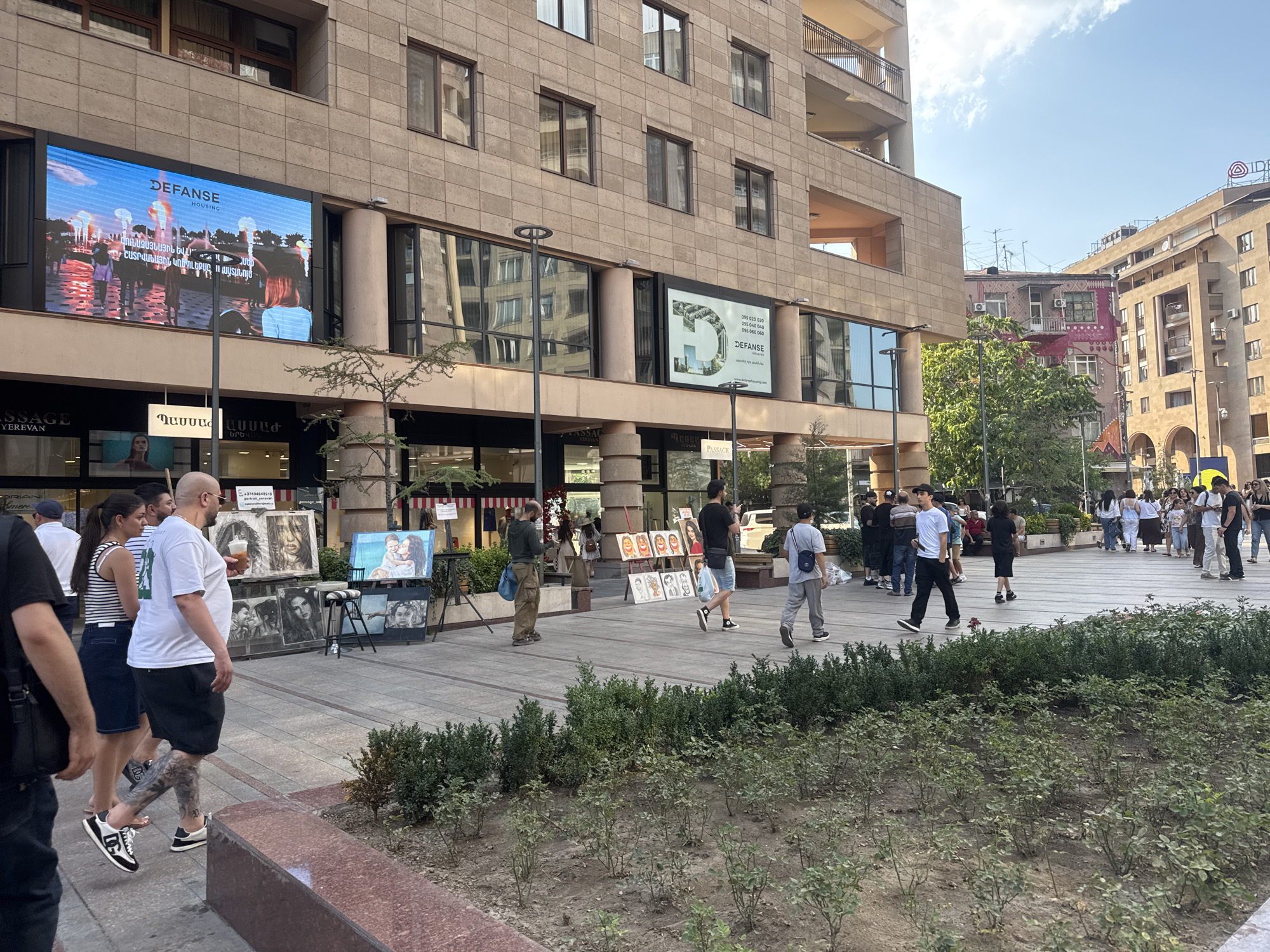 The image depicts a street scene in Yerevan, Armenia, likely in a commercial or pedestrian area. The foreground features a planted area with young, sparse greenery, transitioning to a paved walkway. On the walkway, several artists are displaying their works, which include framed portraits and paintings. People are walking along the paved area, some pausing to look at the art. In the background, a multi-story building with stone facade and numerous windows dominates the scene. Large digital billboards are mounted on the building's exterior, displaying advertisements. Further in the background, other buildings and a street are visible, under a bright, partly cloudy sky. The overall impression is of a lively urban environment with elements of commerce and street art.
