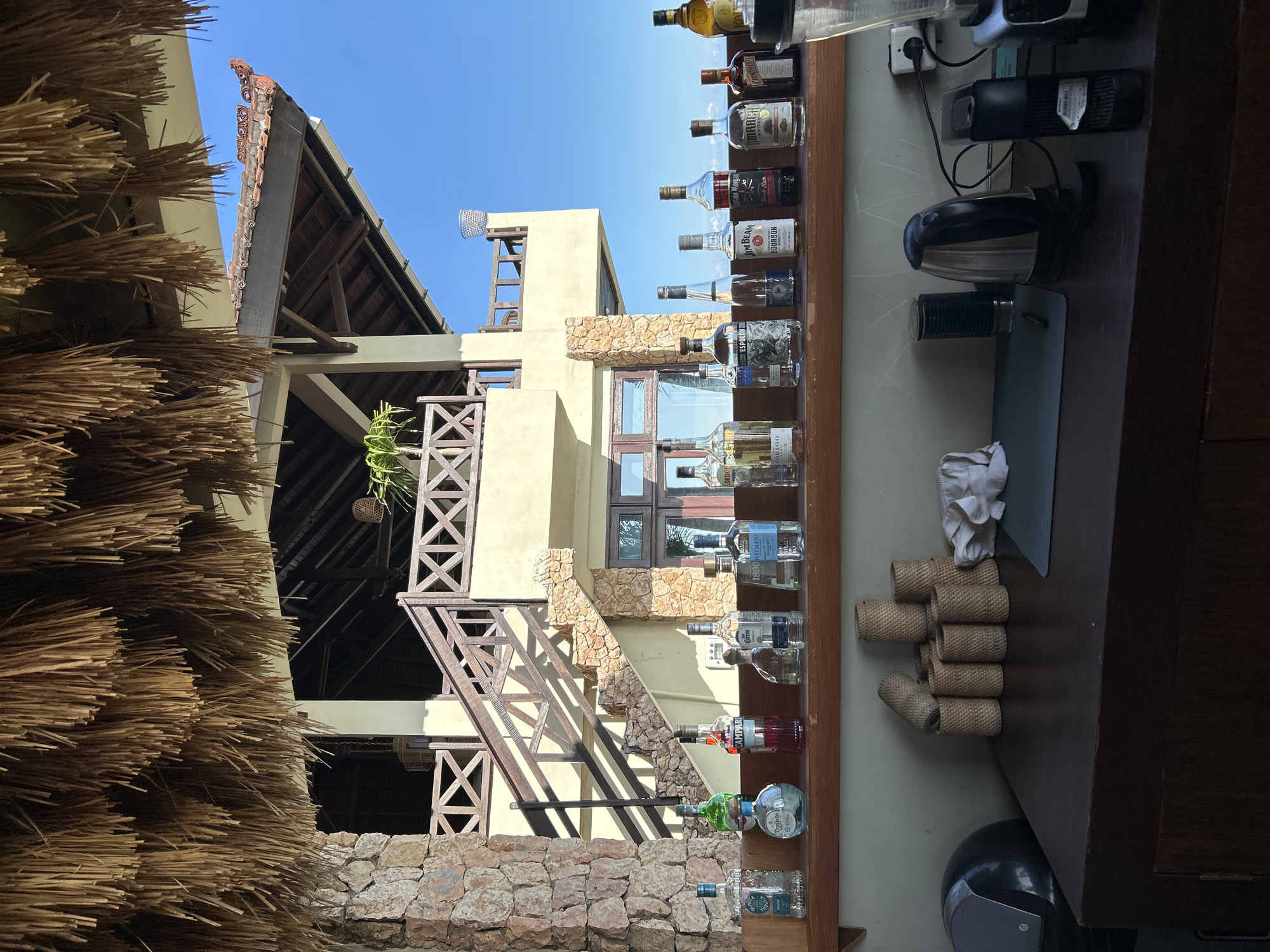 The image captures a bar setup in Senggigi, Indonesia, against the backdrop of a multi-level building under a clear sky.
On the right side, the bar area displays a shelf with various alcohol bottles arranged in a row. Adjacent to the bottles, there are rolls of woven material and a coffee maker. Electrical sockets and equipment cords are visible on the wall behind the bar.
In the background, a building constructed with stone and pale yellow plaster features a wooden staircase with a cross-pattern railing, climbing along the exterior. A thatched roof structure stands to the left of the building, with its straw-like material filling the left foreground. The building's architecture suggests a tropical resort or restaurant setting. The sunlight indicates a daytime scene.