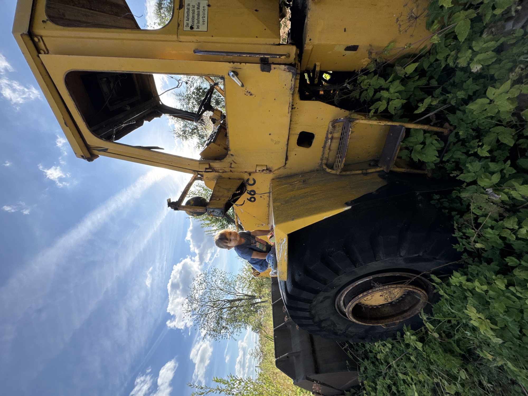 In Innolovo, Russia, a young child sits atop a large, yellow, weathered construction vehicle, possibly a loader, overgrown with green foliage. The child, wearing a blue t-shirt and grey Crocs, is facing left, looking down. The vehicle's cab is open, revealing a steering wheel and exposed wiring. A sign with German text is affixed to the side of the vehicle. The scene is set outdoors under a bright blue sky with scattered white clouds and wispy cirrus formations. The ground is covered in lush green plants.