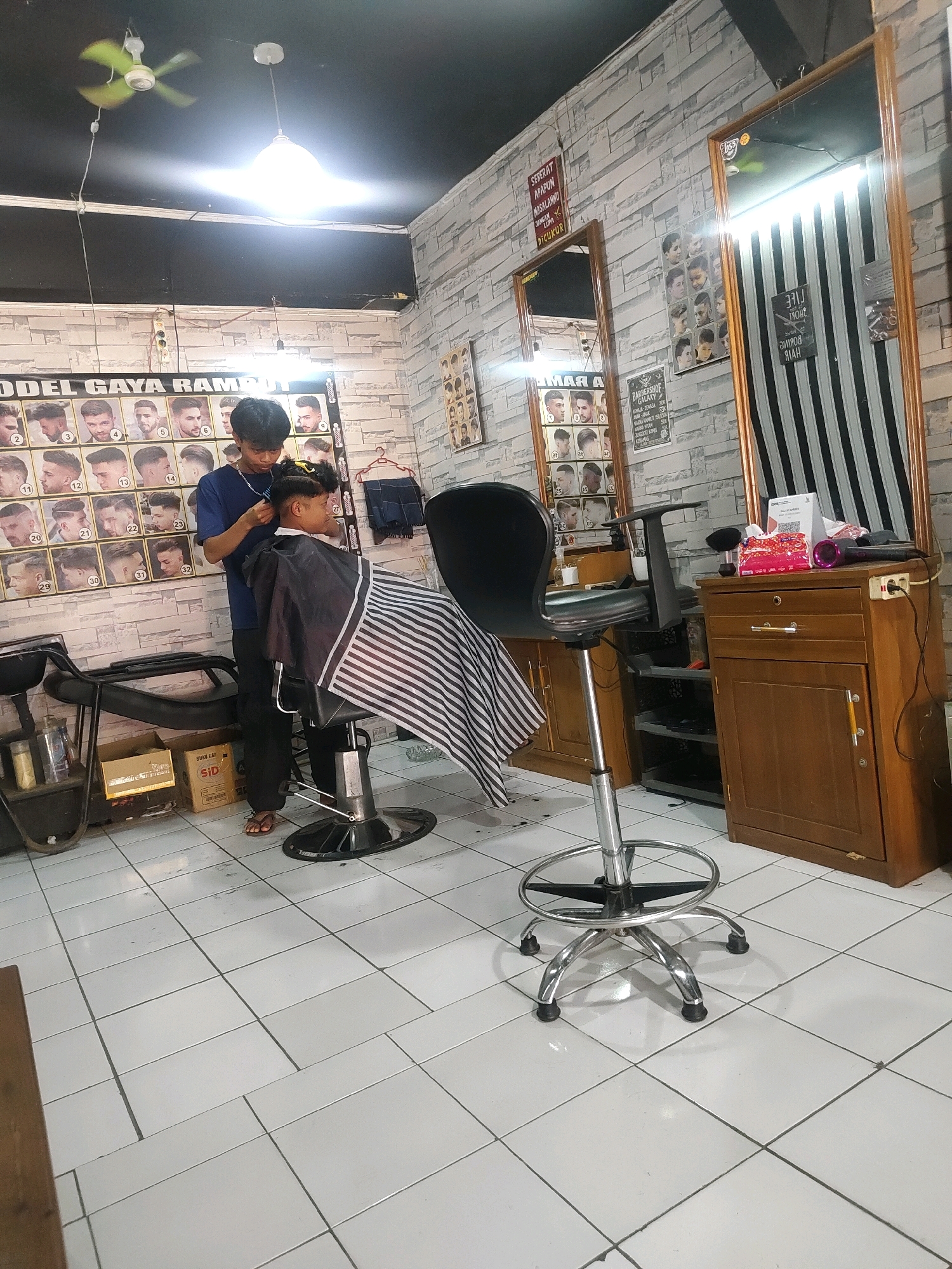 A barber is cutting the hair of a customer inside a brightly lit barbershop located in Sukanagara, Indonesia. The barbershop features a white tiled floor and walls adorned with posters of various hairstyles and a brick-like pattern. A barber's chair with a black seat and chrome base is positioned in the center of the room. A barber, wearing a blue shirt and black pants, is actively cutting the hair of a customer seated in the chair, who is covered with a black and white striped cape. There are mirrors on the walls, and a wooden cabinet with drawers and doors stands to the right. A fan is visible on the ceiling, and various items like boxes, bottles, and a hairdryer are scattered around the shop.