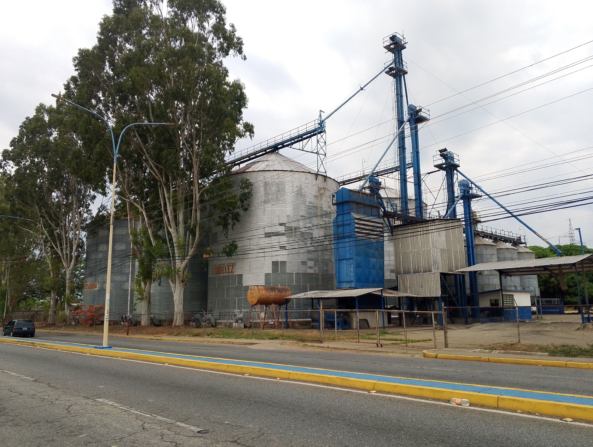 The image depicts a grain storage and processing facility located in San Carlos, Venezuela. The facility consists of several large, corrugated metal silos, connected by a network of blue-painted metal conveyors, towers, and processing structures. A rusted, elevated tank is also visible. The silos are labelled "MARAVILLA" in large letters.
In the foreground, there is a road with a yellow curb and a blue stripe. A dark-colored car is parked on the side of the road. Along the road, there are trees. The sky is overcast and grey. The general environment appears somewhat rundown.