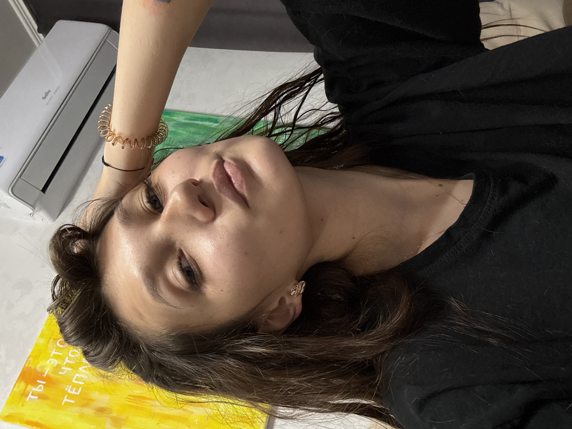 A young woman is captured in a selfie, her face angled towards the camera with her right hand resting on her head. She has dark hair, which is slightly messy, and is wearing a black t-shirt. She is also wearing a gold-toned knot-shaped earring in her visible ear and a spiral-shaped bracelet on her right wrist. In the background, a yellow painting with Russian text and a green painting are visible. An air conditioning unit is mounted on the wall above her. The image appears to be taken indoors, likely in a residential setting. There is no information in the image to indicate the specific location within Novokuznetsk, Russia.