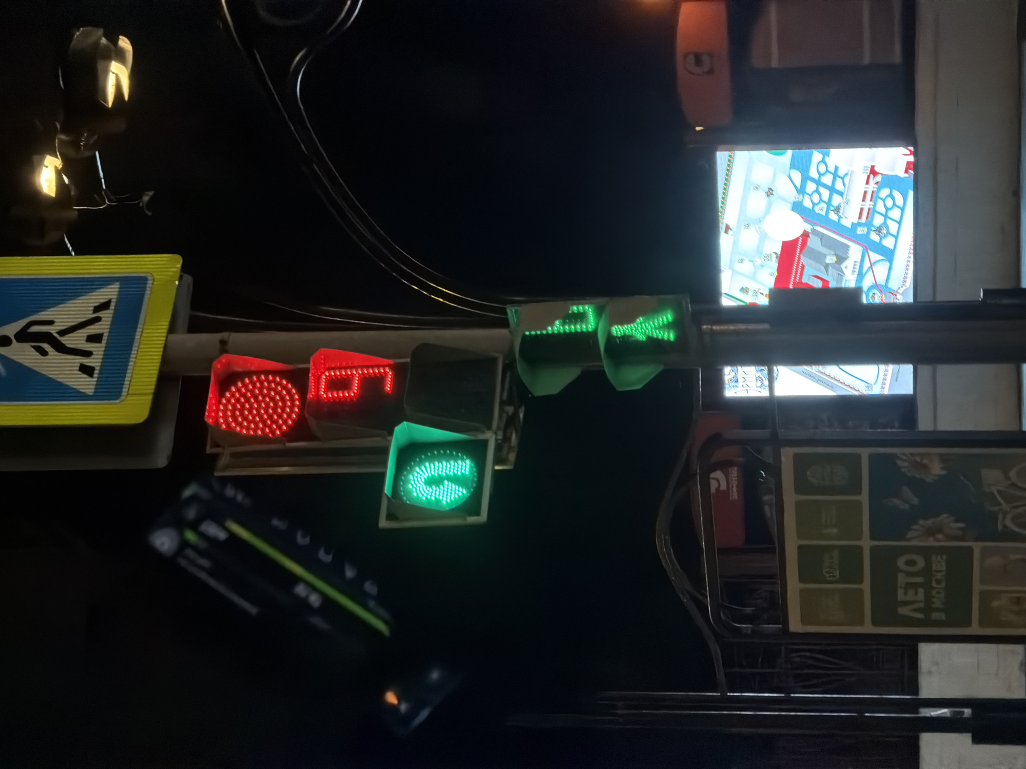 The image shows a street scene at night in Moscow, Russia. A pedestrian crossing sign is visible on the left. A traffic light is lit with a red light for vehicles and a green light for pedestrians. There is a sign that says "Moscow Summer" in Russian. There is another sign with a street map. The scene is illuminated by streetlights.