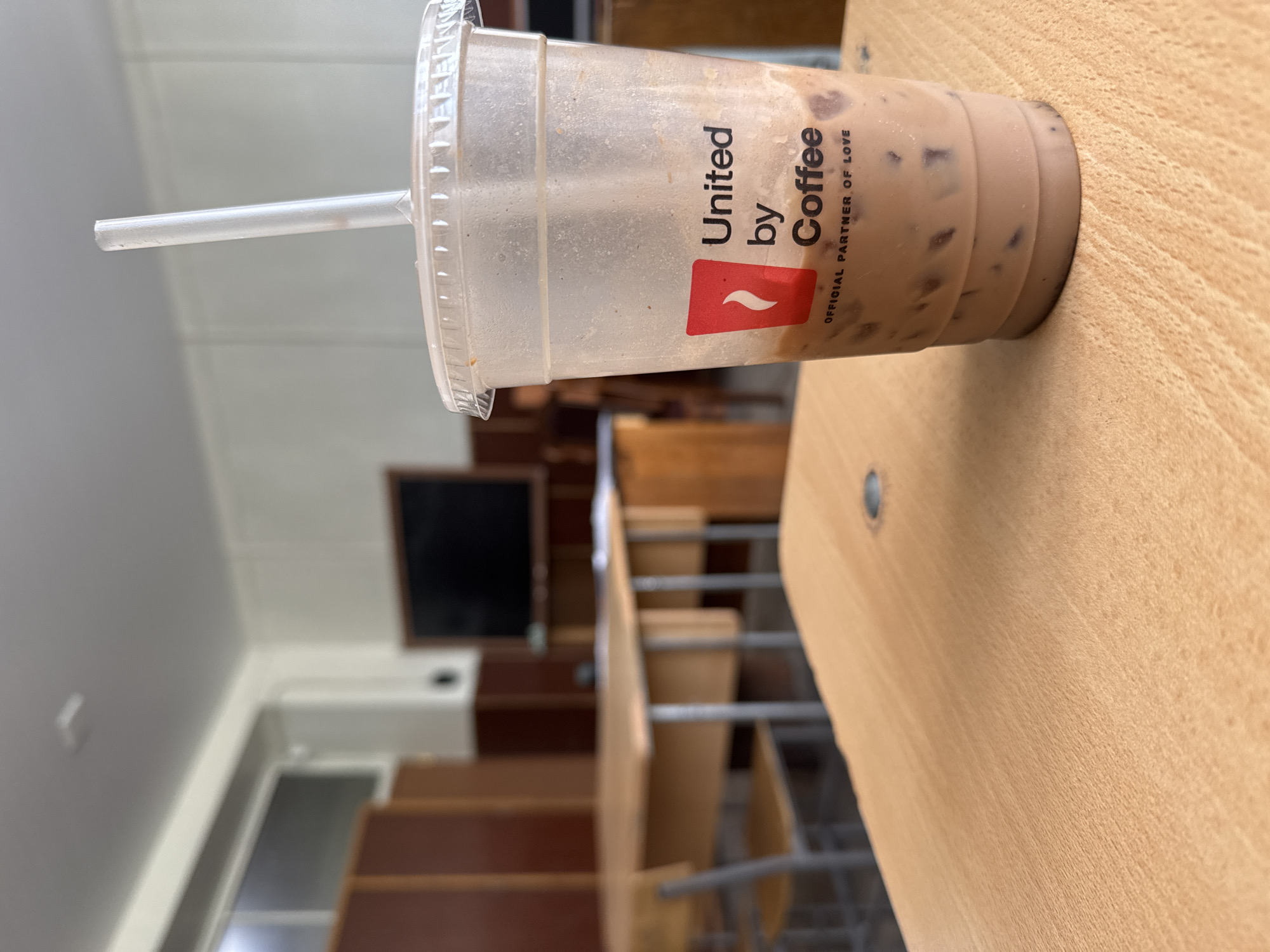 The image displays a clear plastic cup with a straw, containing an iced beverage, resting on a light wooden desk. The cup is partially consumed, approximately one-third full, and shows visible ice cubes in a light brown, creamy liquid, likely an iced coffee or chocolate drink. The exterior of the cup has some condensation and residue.
Prominently featured on the cup is a black and red logo. The text reads "United by Coffee" in black, stacked over a red stylized wave or flame icon, which is positioned next to the word "Coffee." Below this, in smaller black text, it says "OFFICIAL PARTNER OF LOVE." This branding is characteristic of Tim Hortons.
The wooden desk where the cup sits has a visible grain and a smooth finish. In the background, the scene is blurred but reveals an indoor setting, likely a classroom or lecture hall. Rows of light brown wooden desks and chairs are discernible, along with what appears to be a blackboard or monitor on the far wall. The overall ambiance suggests a daytime setting within an educational institution or a similar public space. No people are visible.
Given the location context of Yerevan, Armenia, the presence of a Tim Hortons cup indicates a widespread international brand presence. The image captures a moment of refreshment, possibly during a break or a study session.