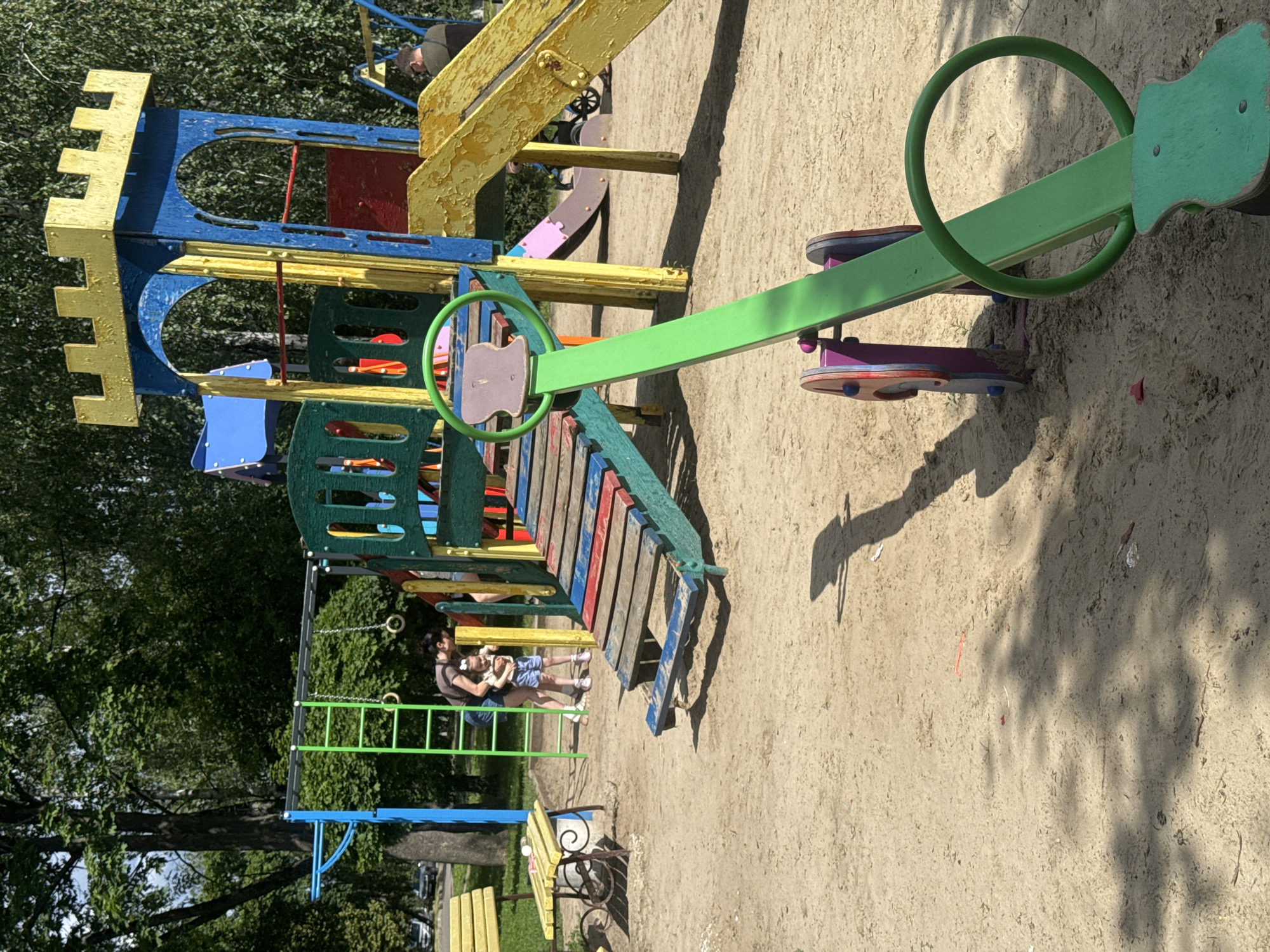 In Poltava, Ukraine, a vibrant children's playground is captured on a sunny day. The scene features a brightly colored play structure resembling a castle, complete with slides and climbing elements. In the foreground, a green seesaw with a circular handle stands on the sandy ground. Nearby, a woman in a dark shirt and shorts embraces a young child wearing a light-colored top and blue shorts. The background is filled with lush green trees, indicating a park or recreational area. A yellow bench and a table are visible on the left, and a glimpse of cars on a street can be seen through the trees. The sand-covered ground is marked with shadows from the playground equipment and trees, suggesting midday sun.