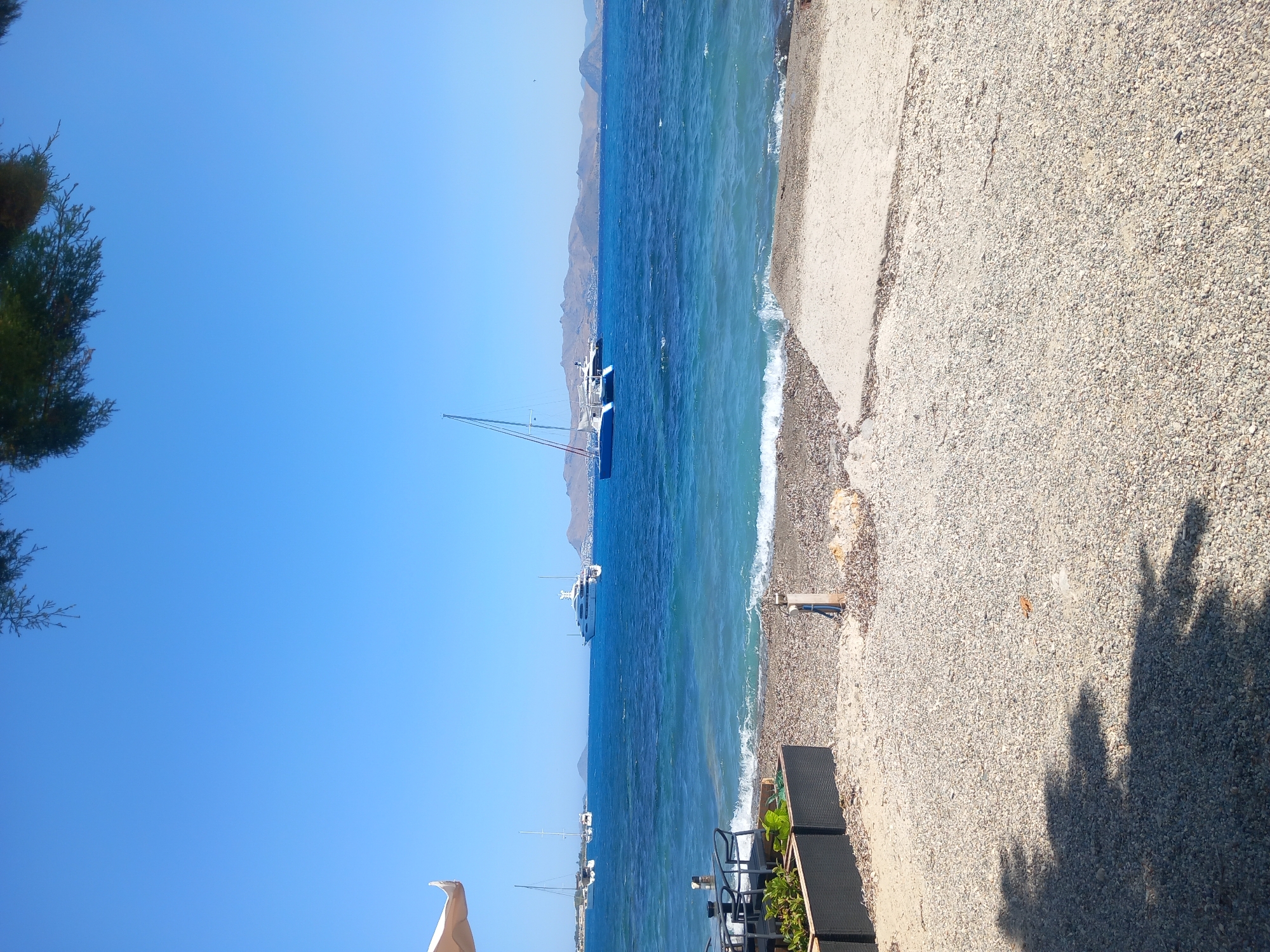 The image shows a bright, sunny day by the sea, likely in the city of Kos, Greece, as suggested by the style of the boats and the coastal landscape. The foreground is a pebbly beach with a concrete slab. To the left, there are dark planters with greenery and some outdoor seating with tables and chairs, partially obscured by a light-colored umbrella. The sea is a vibrant blue with moderate waves. Several boats are visible in the water, including a large white yacht, a blue catamaran with a sail, and other sailboats in the distance. In the background, a chain of islands or coastal mountains are visible under a clear blue sky. The overall impression is a serene and picturesque seaside scene.