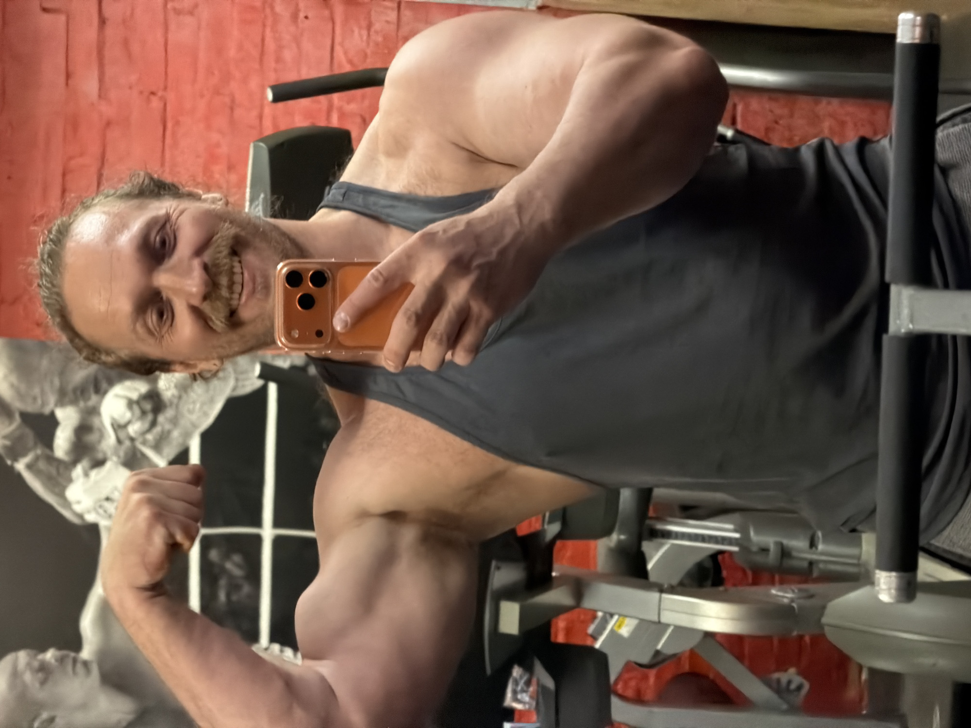 A man in a gray tank top is taking a selfie in what appears to be a gym setting. He's flexing his arm. Behind him, a wall features a black and white mural. The background suggests a fitness environment, likely in a gym. There's no specific information within the image to confirm it is located in the city of Saint Petersburg, Russia.