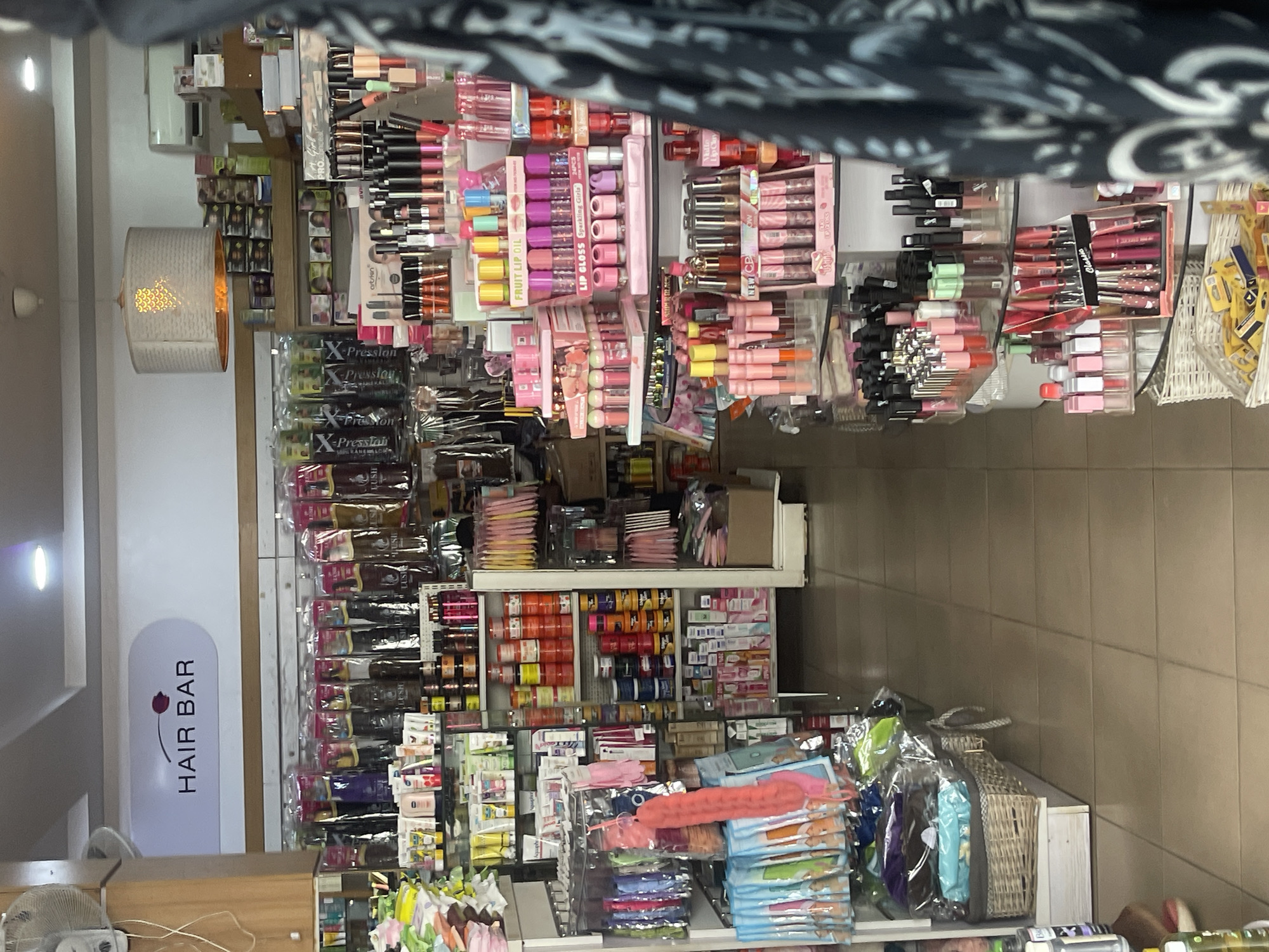 The image shows the interior of a "HAIR BAR" retail store, likely in Abuja, Nigeria, given the products and brand names visible, such as "X-Pression" hair extensions. The shelves are densely packed with a variety of beauty and hair products, including lip gloss, cosmetics, hair extensions, and accessories. The products are arranged vertically on the shelves, creating a colorful and crowded display. Overhead lighting illuminates the scene, and the floor is tiled. The image appears to have been taken from a slightly elevated perspective, with some clothing items partially visible in the foreground.