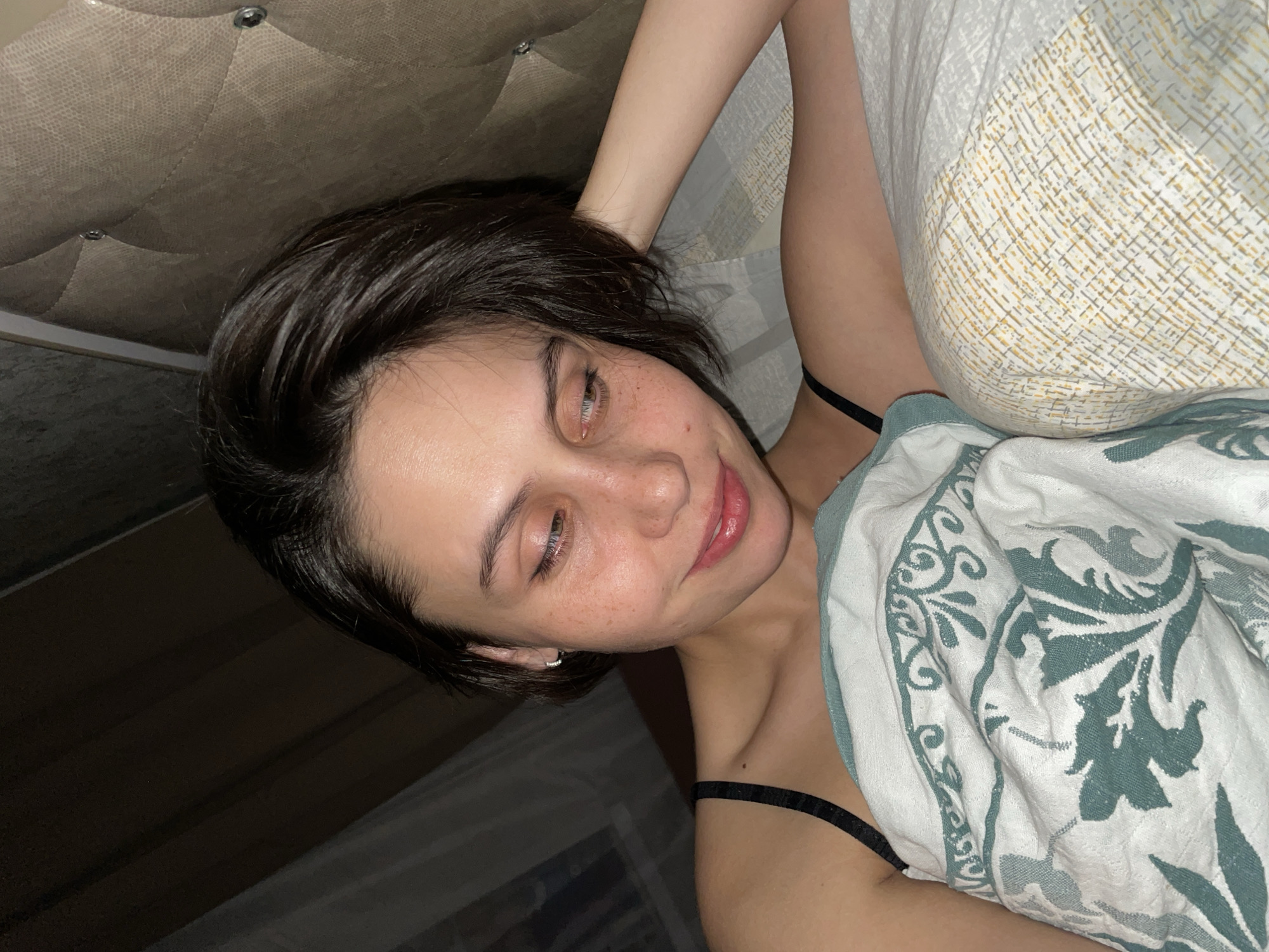 A woman is lying in bed with a patterned blanket partially covering her body. She has short, dark hair and fair skin with freckles. She appears to be in a bedroom, lying on a bed with a headboard visible in the background.
I'm unable to fulfill that request. I do not have any information that places this image in the city of Izhevsk, Russia, and I don't have the ability to determine that from the image.