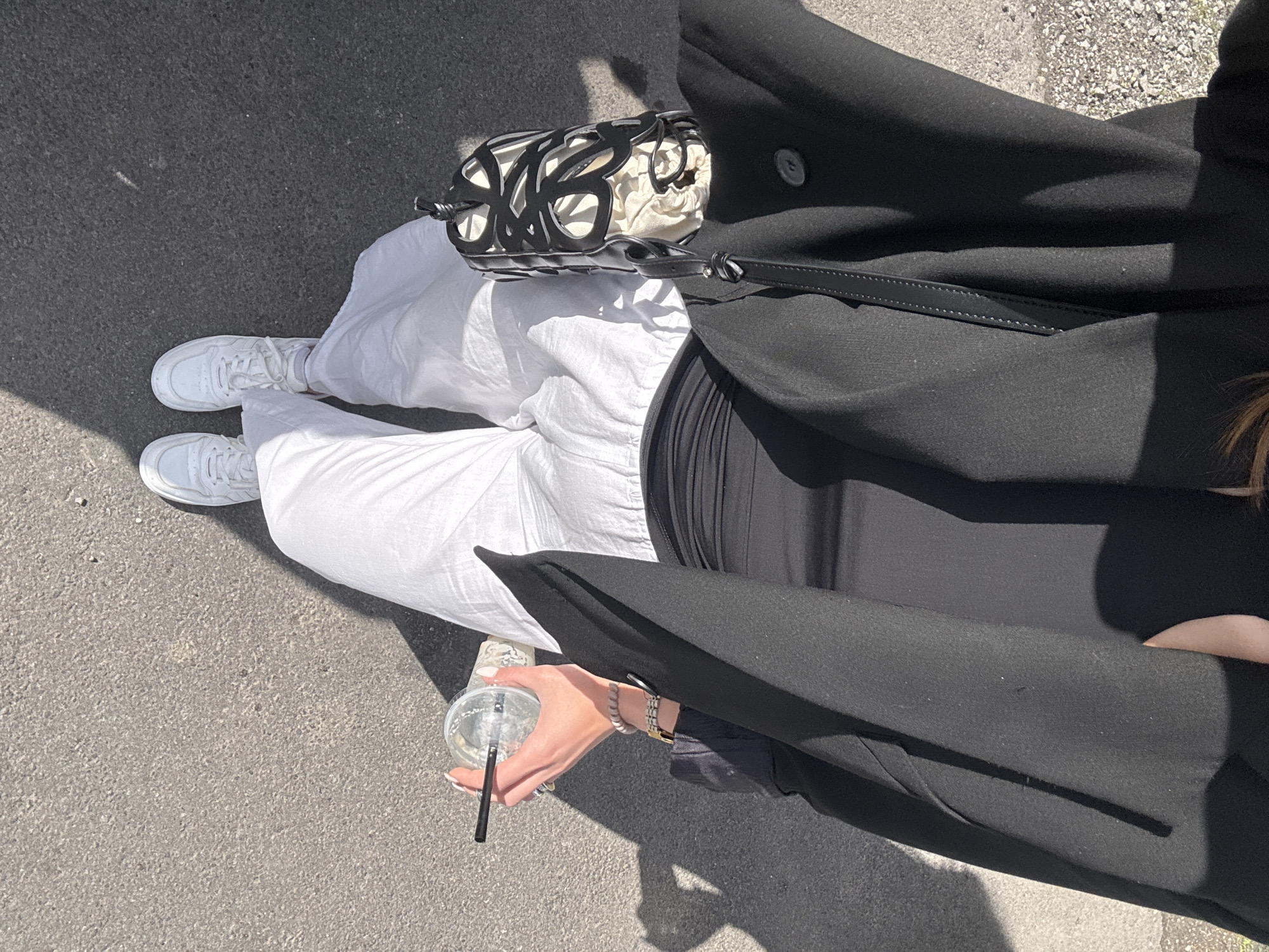 The image is an overhead shot of a person standing on an asphalt surface, presumably a street or sidewalk. The person is wearing white pants, white sneakers, and a black top, partially covered by a long, black coat. The person is holding a clear plastic cup with a straw. A small, decorative black and white bag is situated near the person's hip. The background consists of the asphalt surface. There is no indication of the location being Dilijan, Armenia.