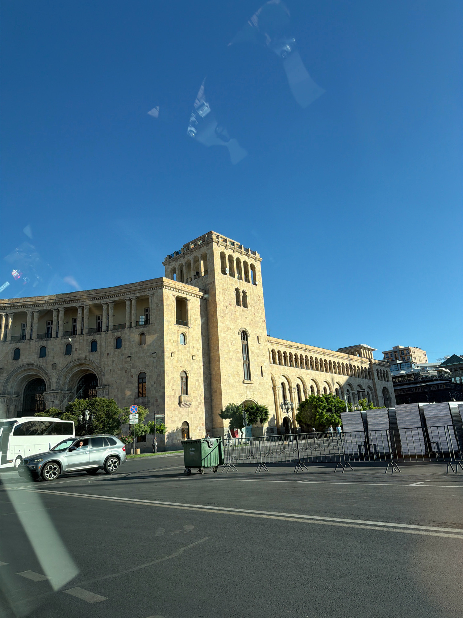 The image depicts Republic Square in Yerevan, Armenia, under a clear blue sky. Dominating the scene is a large, tan stone building with classical architectural features, including arched windows and columns. The building has a prominent tower on its right side and a long, colonnaded section extending to the left. In the foreground, a gray SUV is driving on a paved road. A white bus is parked to the left of the SUV. Several metal barriers are set up along the sidewalk in front of the building, with stacks of white boards behind them. A green trash bin on wheels is positioned near the barriers. Trees are visible in front of the building. Reflections of the sky and possibly a person are visible on the surface from which the photo was taken, likely a vehicle window.