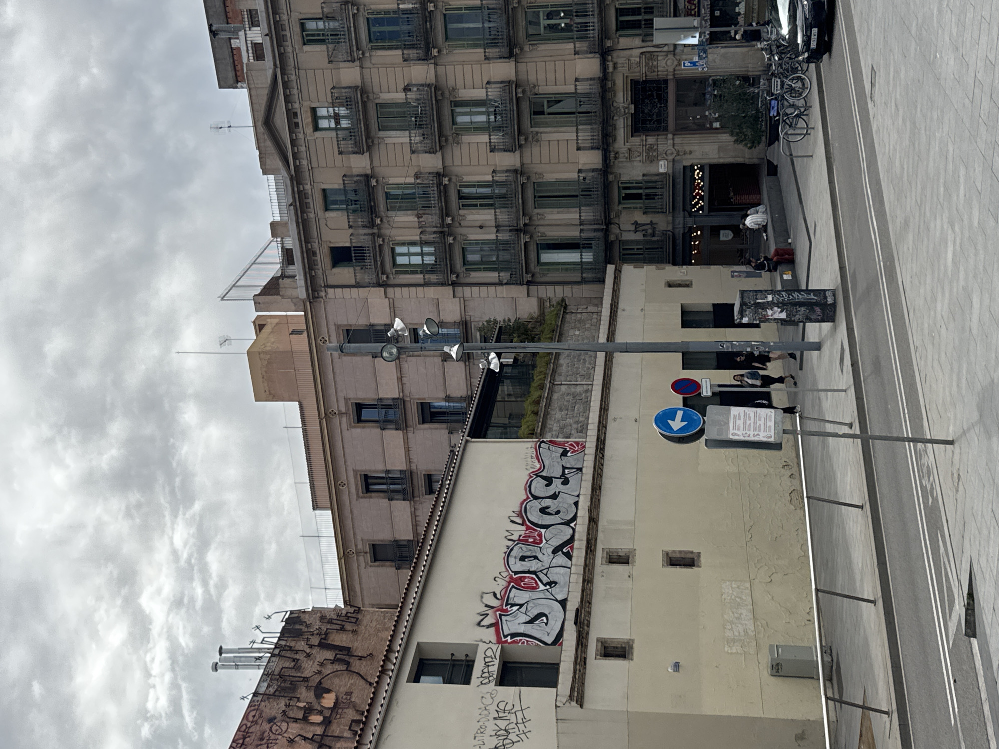 The image captures a street scene in Barcelona, Spain. The street has a sidewalk with pedestrians and a road with white lines. Several buildings line the street, some with graffiti on their walls. A street sign is visible, indicating a direction. There are bicycles parked near a building. The sky is overcast.