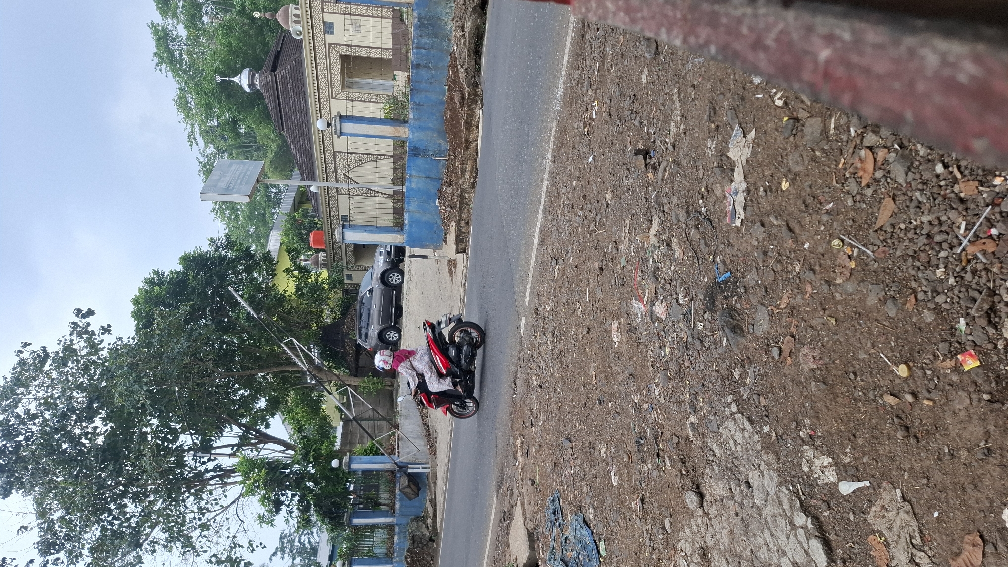 A red motorcycle is being ridden by a person wearing a pink headscarf and a patterned outfit. The motorcycle is on an asphalt road in front of a building with a dome and an Islamic crescent symbol. There are trees and a gray car in the background. The ground in the foreground is covered in dirt and debris. The image was taken in Cilaku, Indonesia.