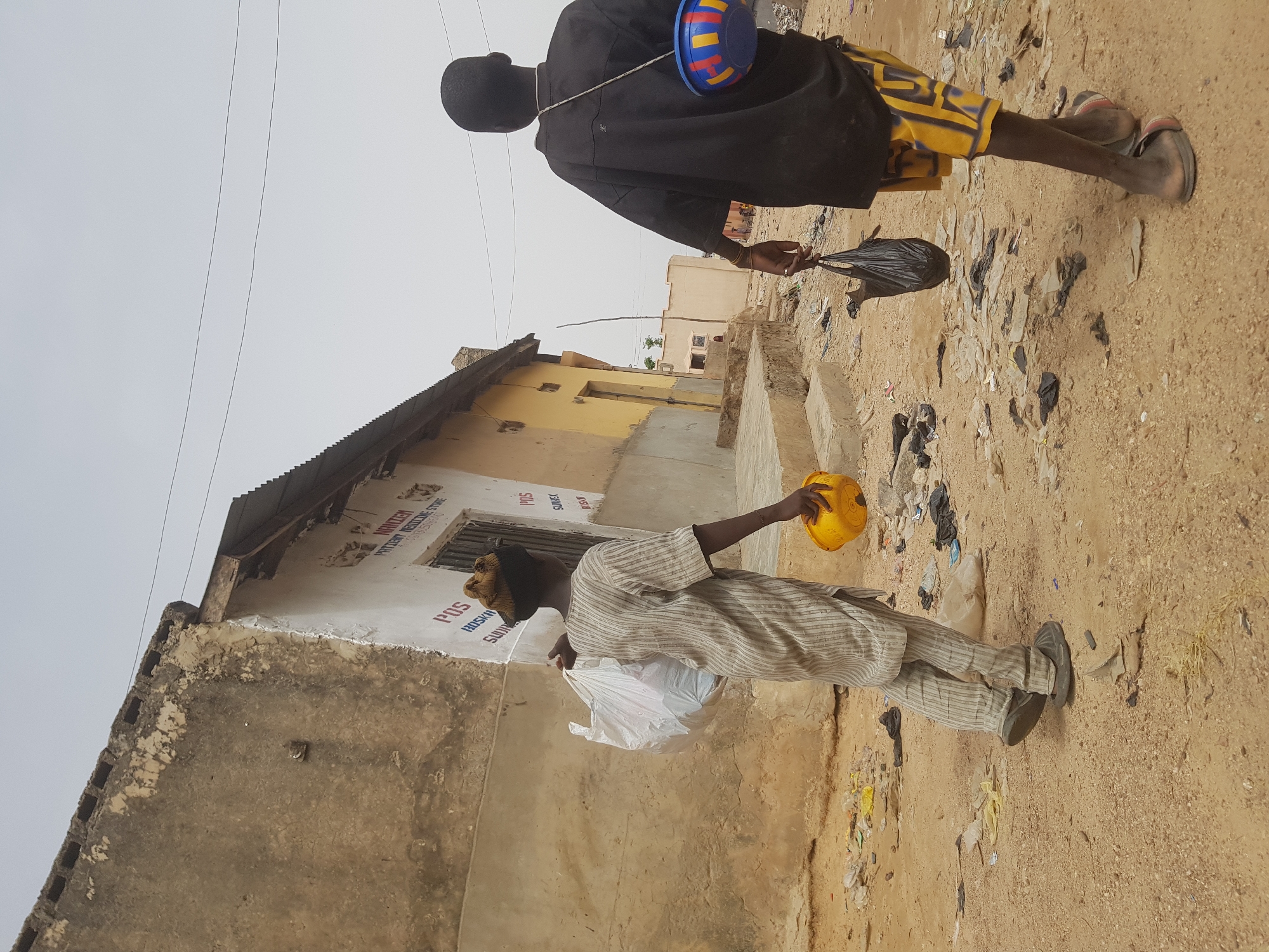 Two individuals are observed on a dirt road littered with debris, presumably in Dutsinma B, Nigeria. The first individual, seen from the back, is wearing a dark garment and yellow shorts, with a blue and red bowl slung across their shoulder and carrying a black plastic bag. The second individual, closer to the foreground, is wearing a striped outfit and carries a white plastic bag and a yellow object. They stand in front of a building with peeling paint and what appears to be a sign reading "POS." The environment suggests a modest or impoverished area.