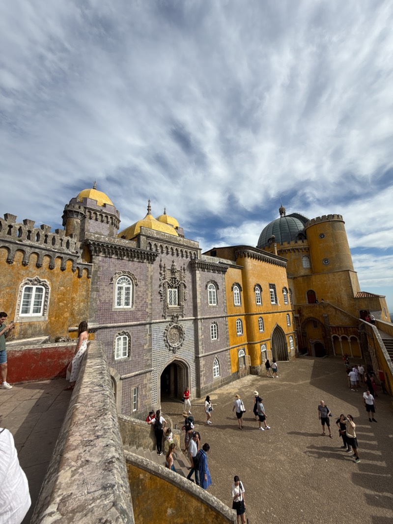 The image shows the courtyard of the Pena Palace, a UNESCO World Heritage Site in Sintra, Portugal. The architecture is colorful and ornate, with yellow, gray, and blue buildings featuring arches, windows, and towers. Several tourists are scattered throughout the courtyard, with some walking, standing, or taking pictures. The sky is partly cloudy, with blue sky visible behind the palace. The courtyard is paved with stones, and a stone wall is visible in the foreground.