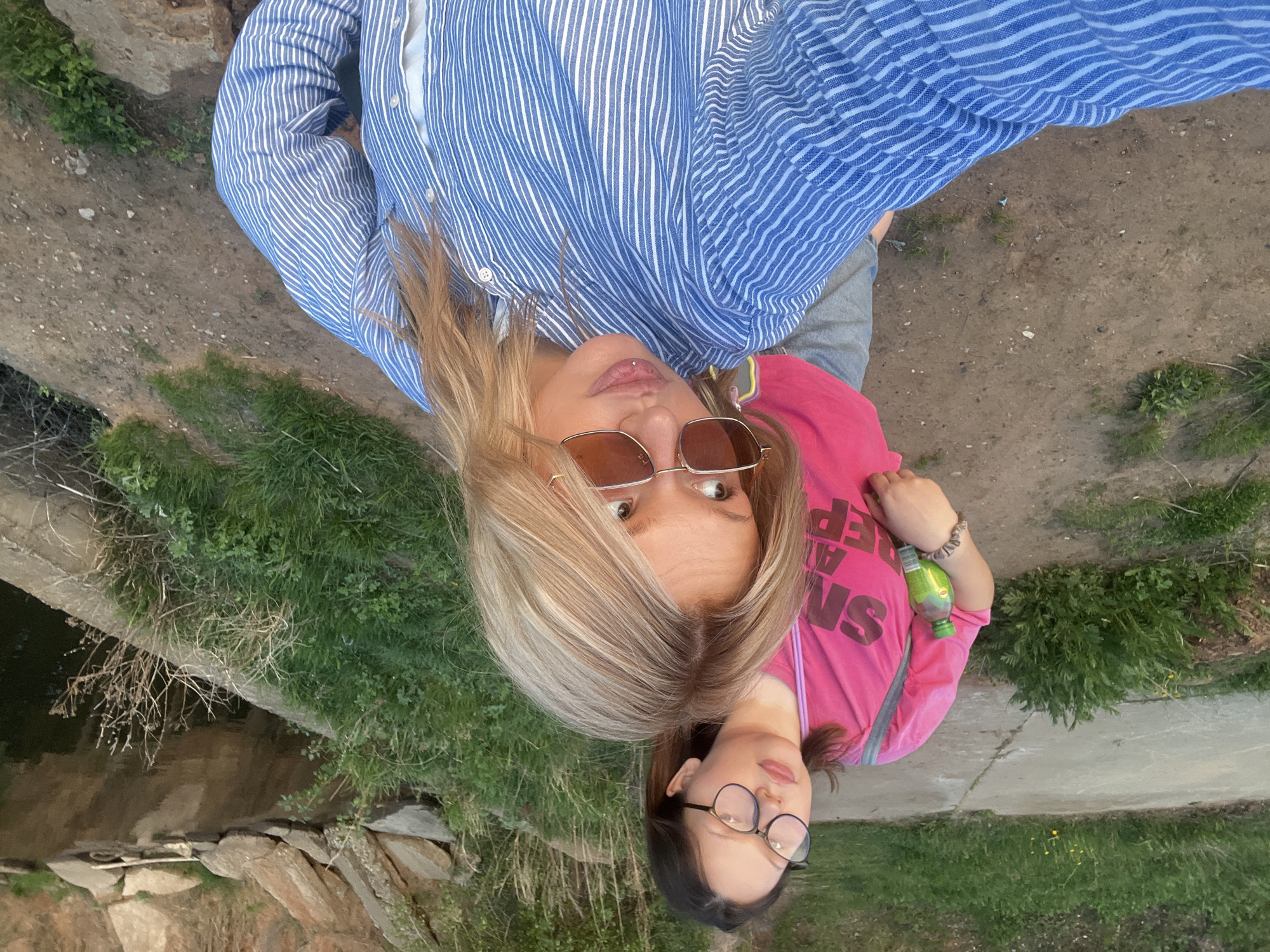 Two females, one blonde and one brunette, are positioned in a selfie frame. The blonde woman is wearing a blue and white striped button-down shirt, sunglasses, and has blonde hair. The brunette, possibly a younger female, wears a pink t-shirt, eyeglasses, and has dark hair. She is holding a green-labeled beverage. They are standing near a body of water with stone banks and vegetation. The background includes concrete and dirt ground with scattered grass. The photo was taken outside in daylight. The city location is Chaikovsky, Russia.