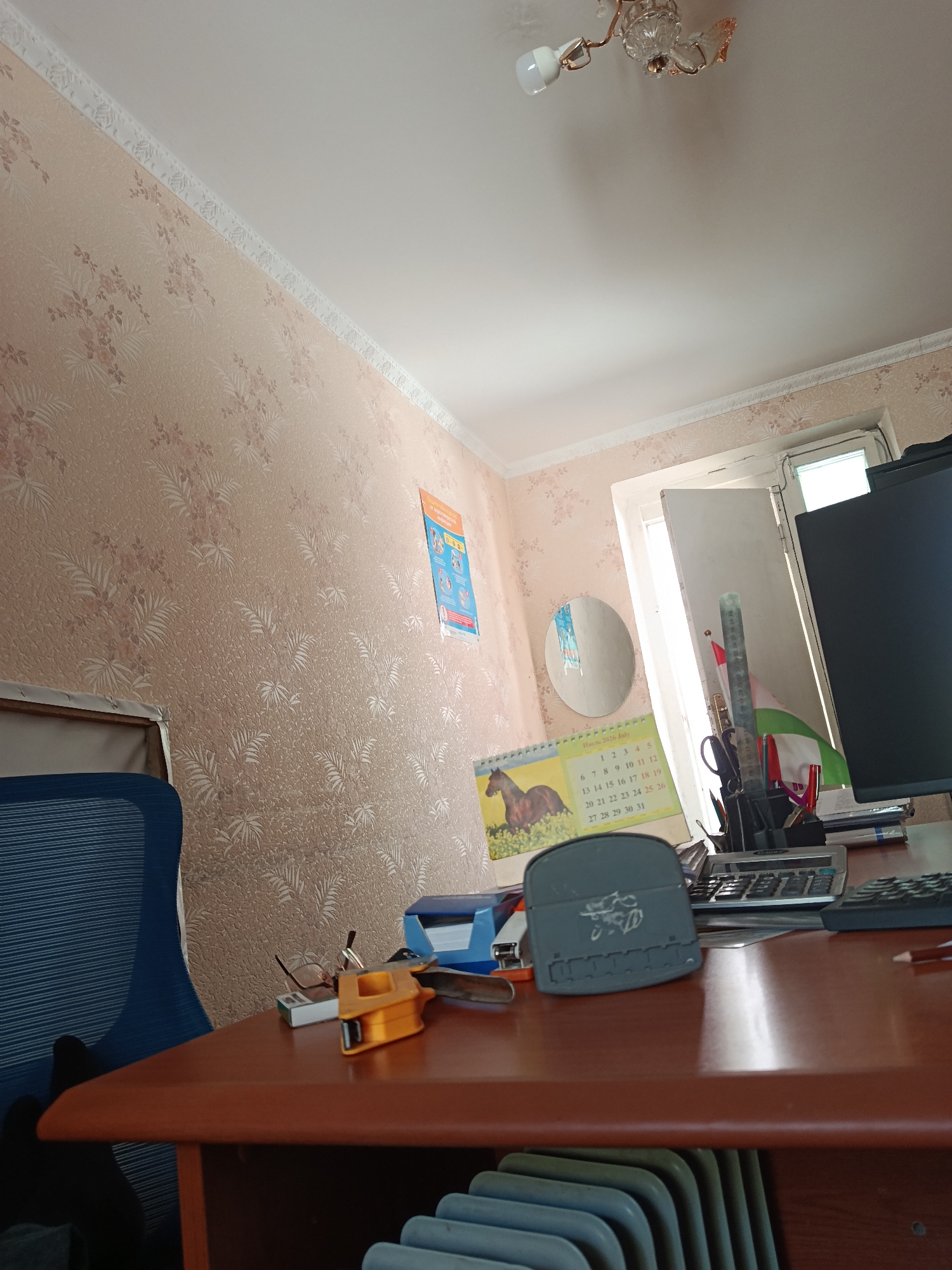 The image shows a desk with various office supplies in a room with light pink floral wallpaper. On the desk, there is a computer monitor, a calculator, a ruler, pens, scissors, a calendar showing the month of July 2026 with a horse on it, and a stapler. A blue office chair is partially visible on the left side of the image. A window is open behind the desk, and a light fixture with an LED bulb is visible on the ceiling. The overall impression is a typical office or workspace.
