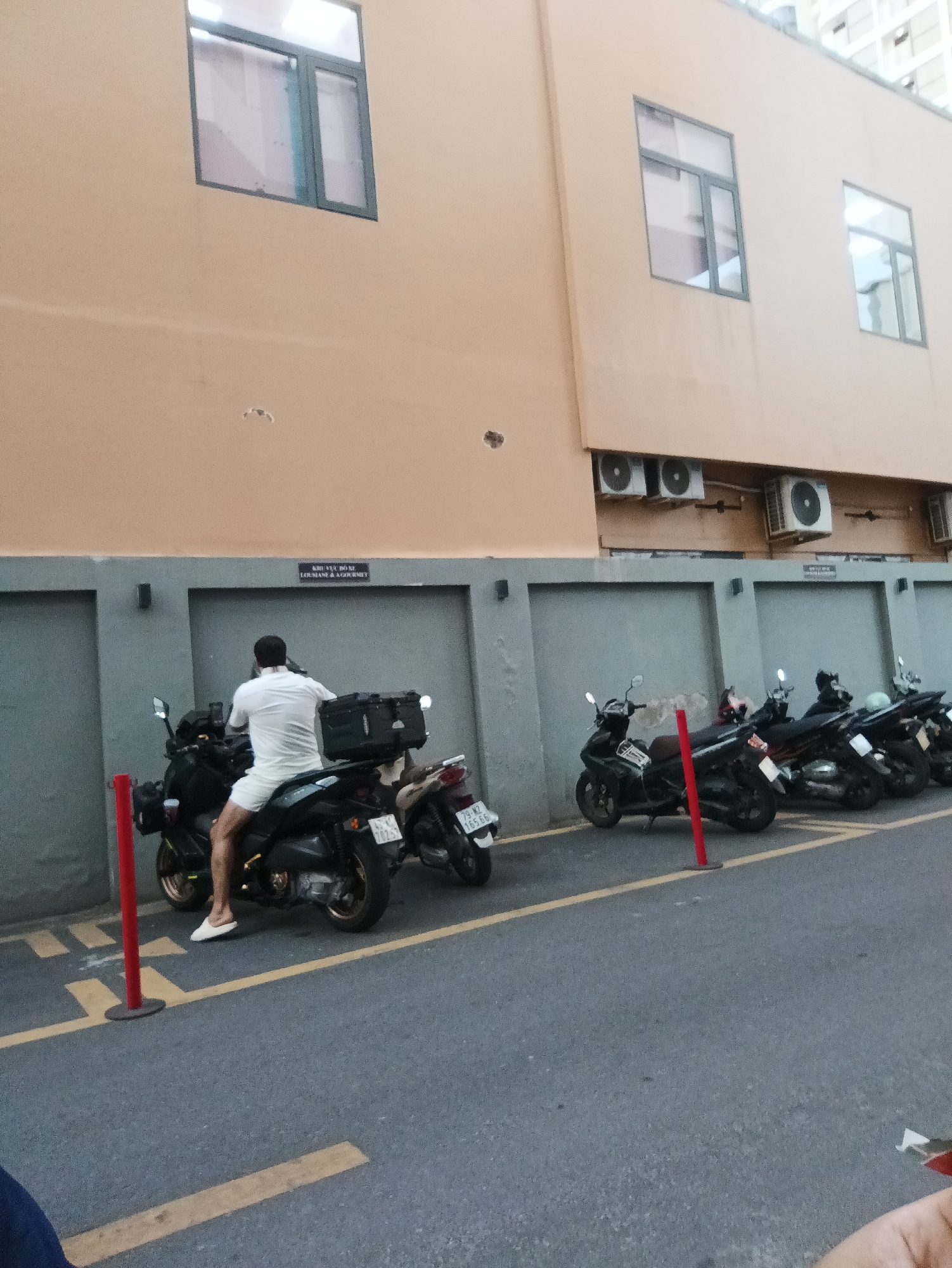 A man wearing a white short-sleeved shirt and white shorts, white flip-flops, is seated on a black motorcycle. The motorcycle has a black cargo box mounted on the rear. The man is facing away from the camera, looking towards a gray wall with several closed garage doors. The motorcycle is parked in a lot with yellow parking lines on the asphalt. Other motorcycles are parked in a row behind his. The building behind the wall is a light orange color and has windows and air conditioning units visible. The scene is likely in Nha Trang, Vietnam, given the style of the motorcycles and the architecture.