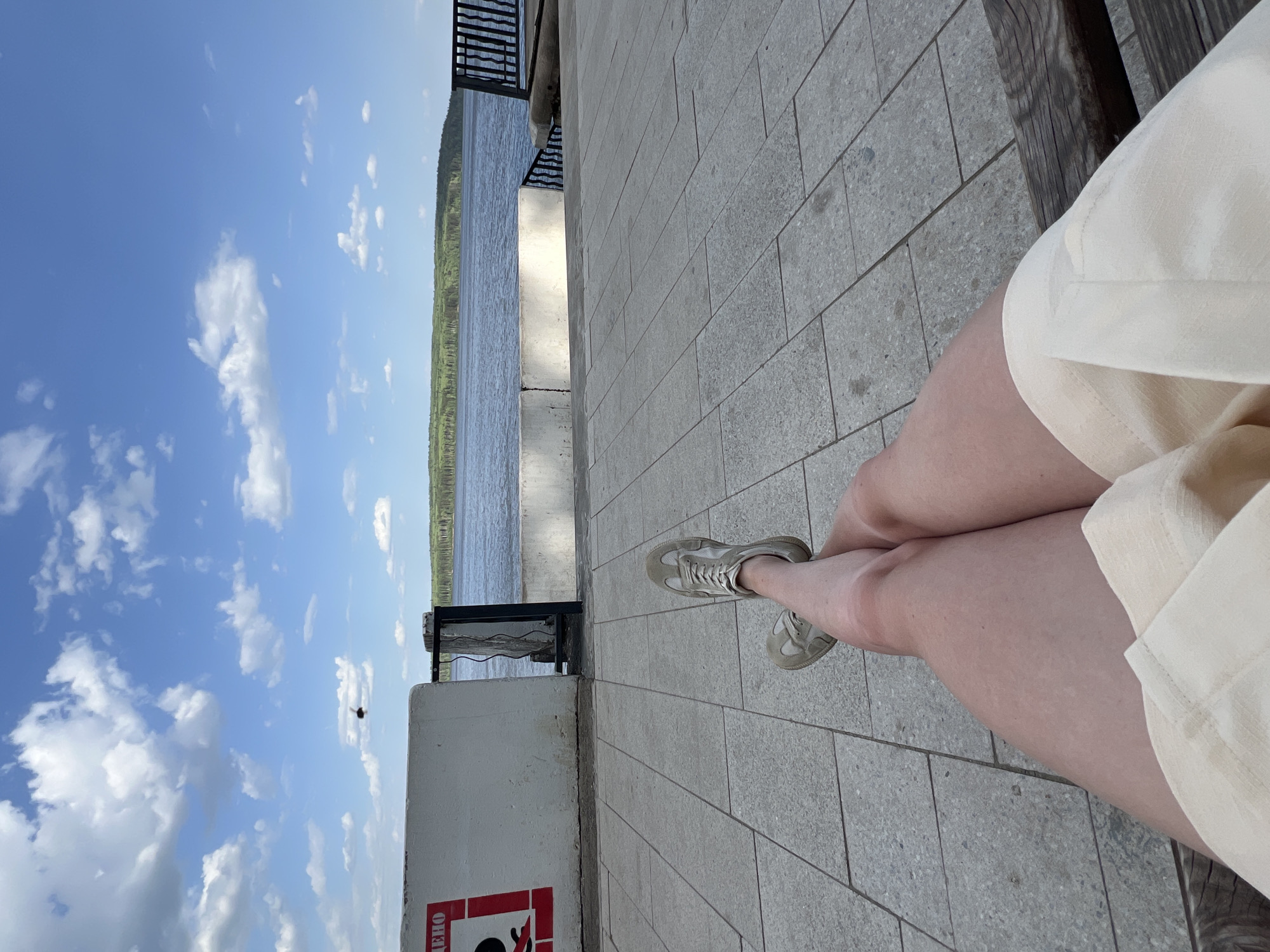 The image shows a person sitting on a bench overlooking a body of water in Chaikovsky, Russia. The person is wearing light-colored shorts and shoes, and their legs are crossed. The scene includes a view of the sky with clouds, a lake or river, and a shoreline with trees. There is a concrete wall or barrier with what appears to be a fire extinguisher sign. The flooring is made of rectangular stone tiles.
