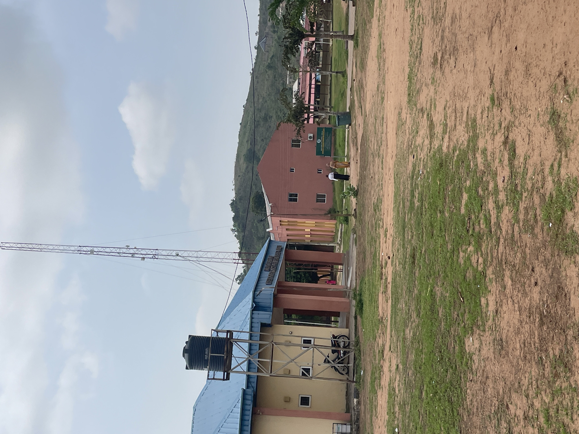 The image depicts the front view of the Kogi State Ministry of Information and Communication in Lokoja, Nigeria. A tall telecommunication tower stands prominently in the background, to the right of a light blue-roofed building. To the left of this building, a black water tank is mounted on a metal frame structure. Adjacent to this, a motorcycle is parked. The building itself has a light yellow facade with a blue roof and red pillars supporting an overhang at the entrance. To the right of the blue-roofed building, a salmon-pink colored building is visible, featuring several windows and an air conditioning unit on its exterior wall. Two individuals are walking away from the camera in front of this pink building, with a green signpost placed between them and a trash can. In the distance, more buildings and palm trees are visible, set against a backdrop of a lush green, hilly landscape. The foreground consists of a patch of grass and bare, sandy earth, with some scattered debris. The sky is overcast with scattered clouds.