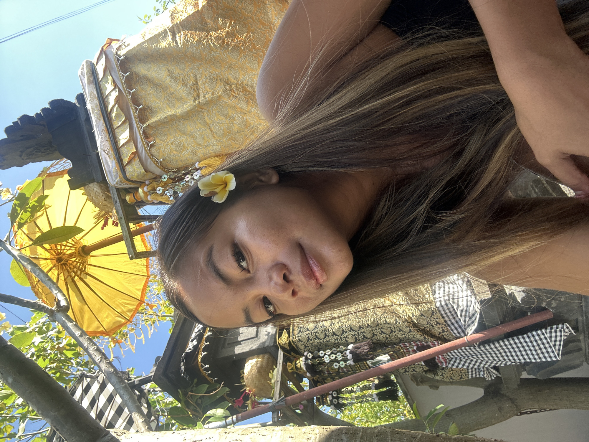 The image shows a young woman with long, dark blonde hair, adorned with a small yellow flower tucked behind her ear. She is looking towards the camera, with a gentle smile and slightly parted lips. Her eyes are open and looking directly at the viewer, conveying a sense of warmth and openness.
The background is a vibrant and somewhat chaotic scene, suggesting an outdoor setting. A large, ornate yellow umbrella with intricate patterns is visible, held aloft by some unseen structure. Elaborate gold and black patterned fabric, possibly part of a traditional garment or decoration, drapes around the scene, interspersed with glimpses of green foliage and a clear blue sky. There are also hints of what appear to be carved wooden elements and a woven basket. The overall impression is one of cultural richness and possibly a festive atmosphere.
The lighting suggests it is daytime, with the sun illuminating the yellow umbrella and casting shadows, indicating a bright, sunny day. The presence of foliage and the style of the decorations strongly suggest a tropical location, consistent with the provided location context of Sukawati, Indonesia. The woman's relaxed posture and pleasant expression suggest she is comfortable and enjoying the moment, perhaps participating in or observing a local event or ceremony. There is no visible text in the image.