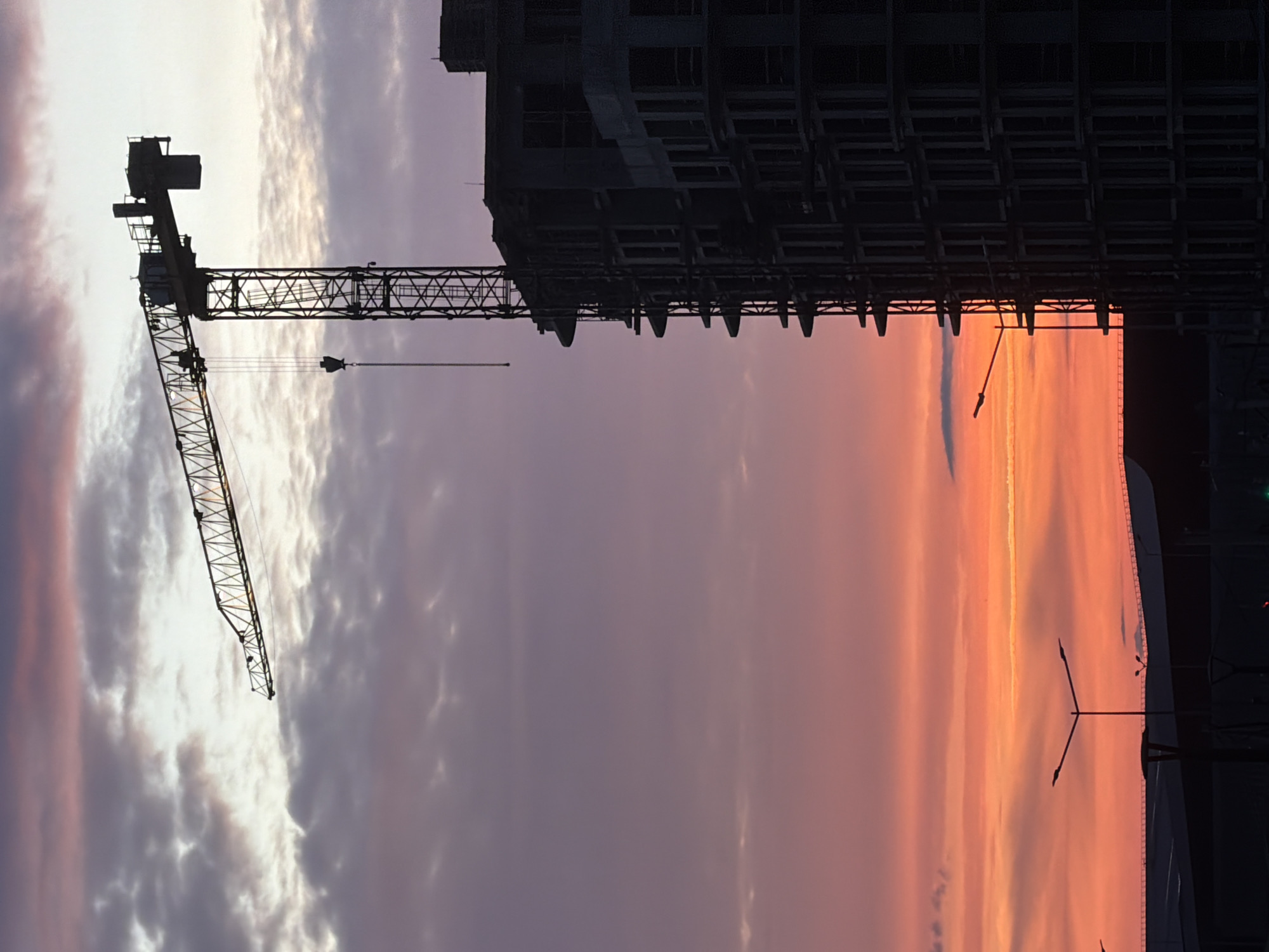 The image depicts a construction site at sunset in Minsk, Belarus. A tower crane, silhouetted in black, stands prominently to the left of a multi-story, unfinished building, also rendered in dark silhouette, occupying the right side of the frame. The crane's horizontal jib extends leftwards, with a hook suspended from a cable. The building displays multiple vacant window apertures and exposed structural elements. The sky exhibits a gradient of colors, transitioning from pale grey and light purple at the top, through shades of violet and pink in the mid-ground, to a vibrant orange and red along the horizon. Various cloud formations are present, appearing diffuse in the upper sky and more defined against the brighter lower sky. In the lower foreground, two streetlights with Y-shaped arms are visible as dark silhouettes, alongside other indistinct structures at ground level. No individuals are visible.