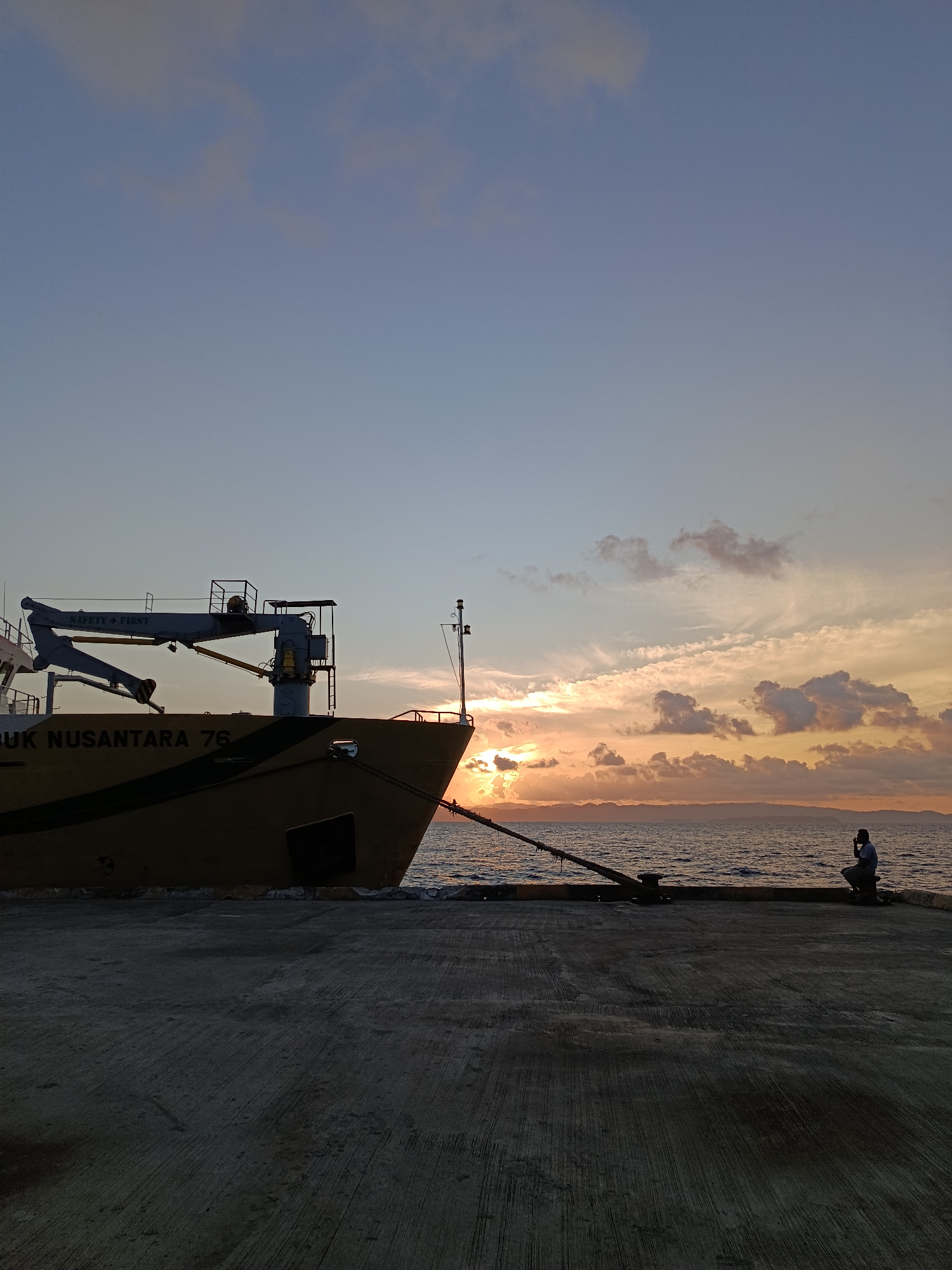 The image depicts a serene sunset scene at the port of Mansalean, Indonesia. A large cargo ship, identified as "RUK NUSANTARA 76," is docked along the pier, its yellow hull contrasting with the twilight sky. A crane extends from the ship, and a person is seen sitting on the pier, looking out at the ocean. The sun is setting, casting a warm glow over the water and clouds. The concrete pier stretches across the foreground, leading towards the ship and the sea. The overall atmosphere is peaceful and tranquil, capturing the end of a day in this coastal Indonesian city.