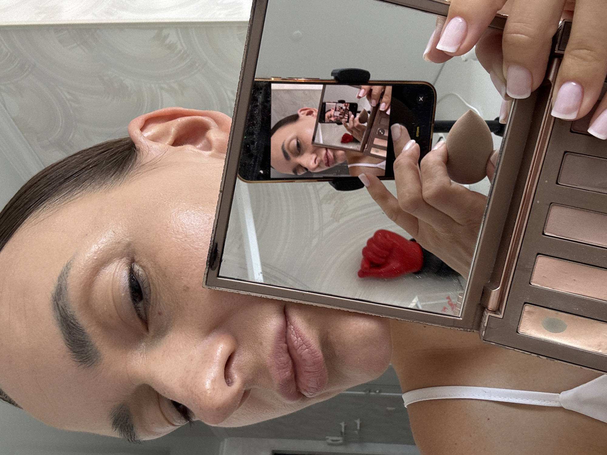 A close-up shot focuses on a woman's face, her left side and lower face are visible as she holds a makeup palette and a smartphone which is angled to reflect her face and another smaller mirror, creating a series of reflections. She is looking at the camera, with her right eye most prominent. Her skin appears smooth and lightly made up. Her hair is pulled back tightly. She is wearing a white strap top. The background is a plain, textured wall with a soft grey and white pattern. There is no indication of the location being Sochi, Russia.