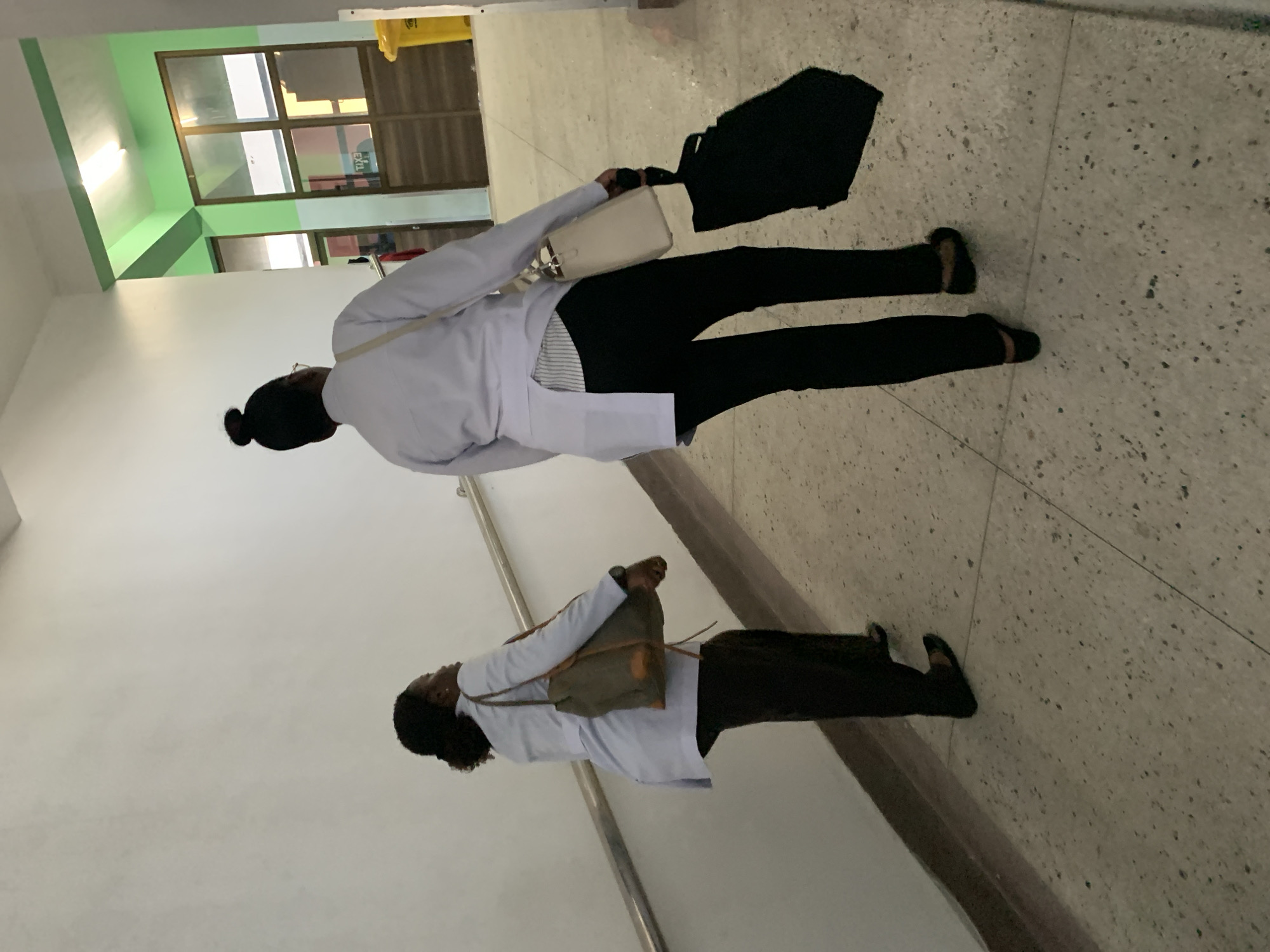 The image shows two individuals, both appearing to be women, walking in an indoor hallway. They are wearing white coats and dark pants. One carries a black bag and a cream-colored purse. The other carries a brown bag. The hallway has a window, and a handrail is visible on the wall. The location is potentially in Dar es-Salaam, Tanzania.