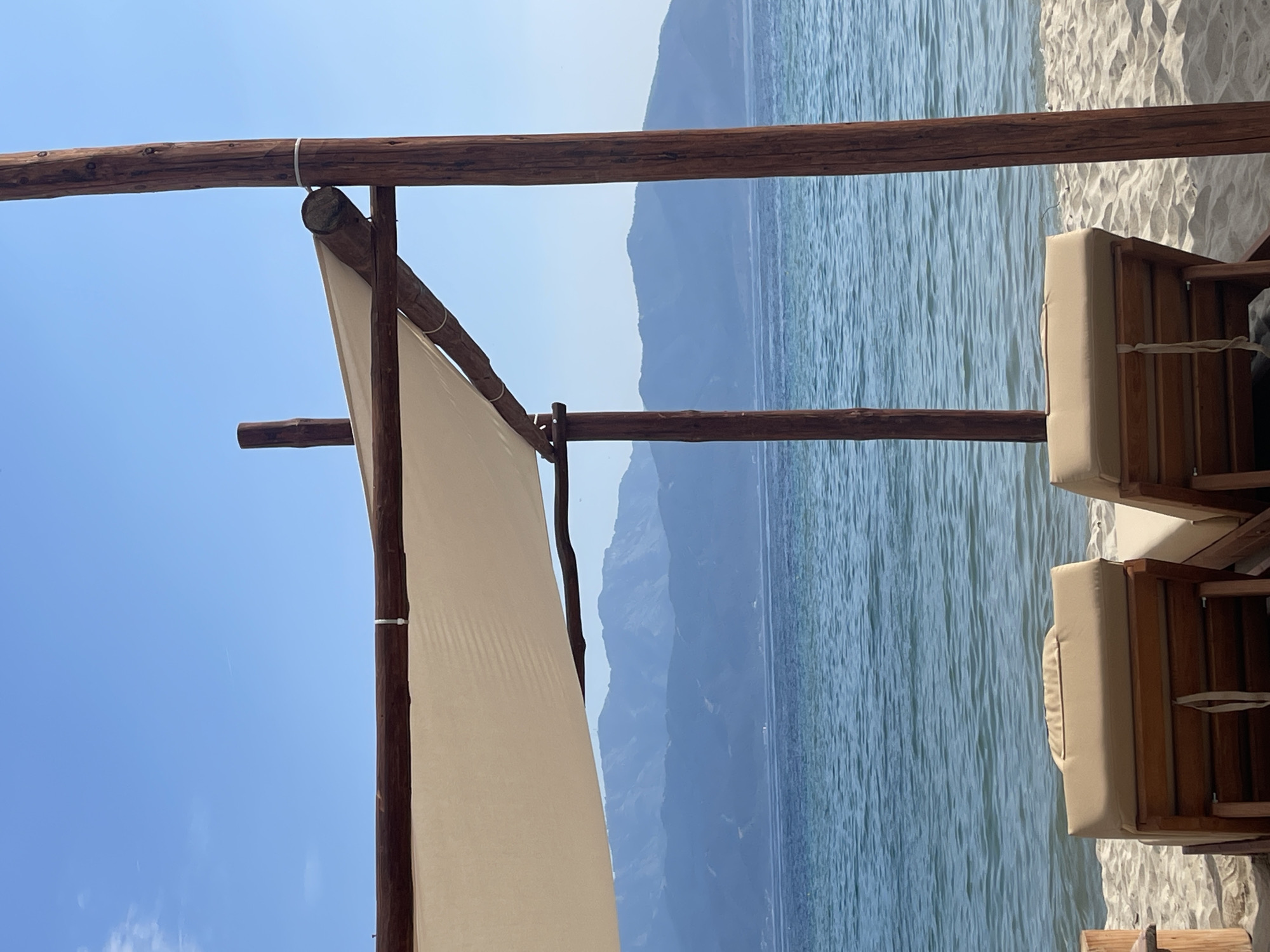 The image captures a serene beach scene in Keramoti, Greece, under a clear, bright blue sky with minimal high-altitude clouds. In the foreground, two empty wooden lounge chairs with light beige cushions rest on light-colored sand, facing the calm, light blue-turquoise sea. The chairs are partially framed by a rustic shade structure composed of dark brown, rough-hewn wooden poles and beams, supporting a taut, light beige fabric canopy. White ties secure elements of this structure. Beyond the water, a range of distant, hazy blue mountains creates a layered backdrop across the horizon. No individuals are present in the scene.