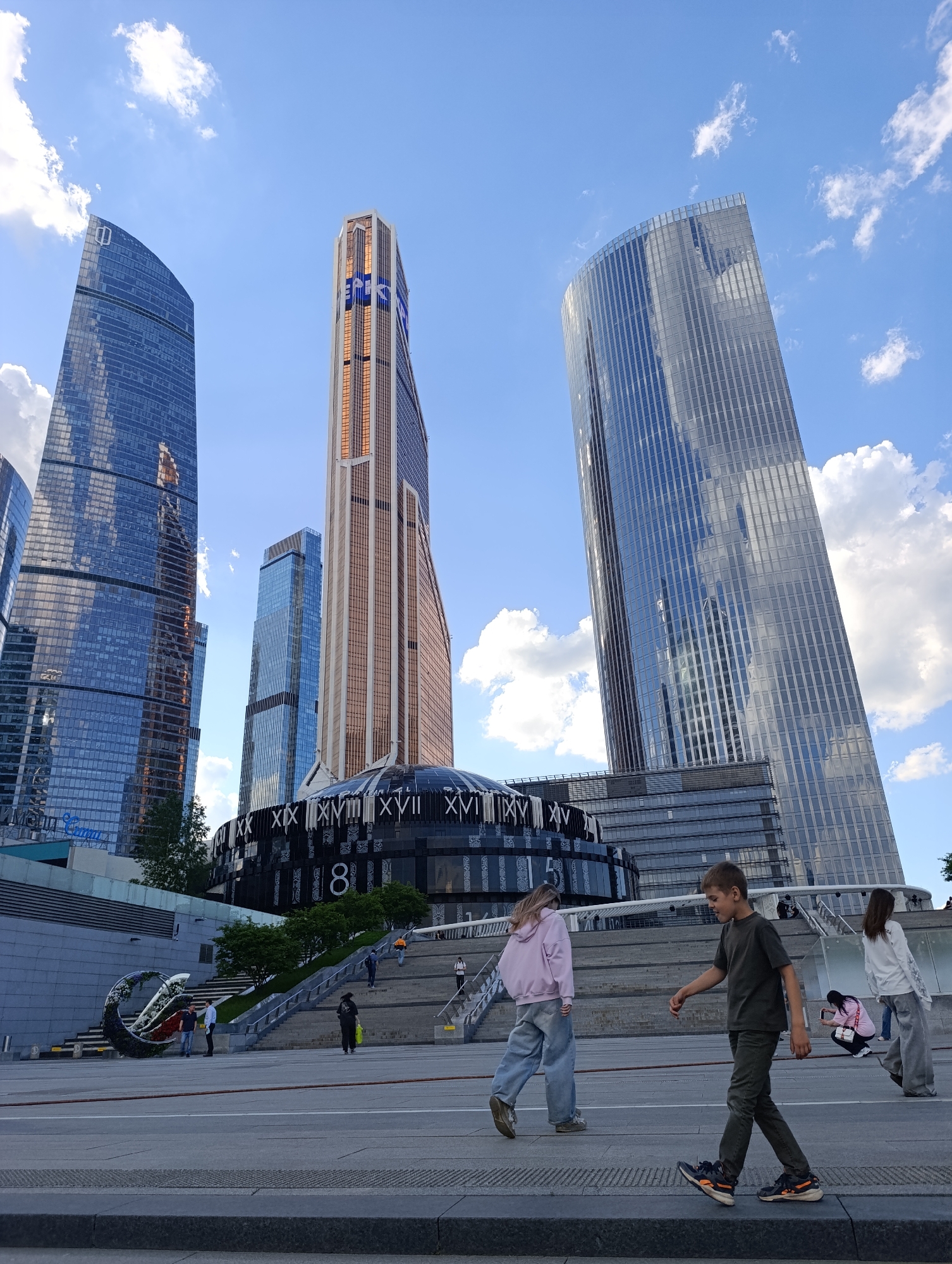 This vertical image captures a modern urban landscape in Moscow, Russia, specifically the Moscow International Business Center (Moscow City), under a bright blue sky with scattered white clouds, suggesting a clear day, possibly late afternoon.
The scene is dominated by several imposing skyscrapers with reflective glass facades. On the far left, a tall, dark blue-tinted glass tower with a subtle round logo near its top stands prominently. To its right, a much taller skyscraper features a distinctive gold and brown patterned exterior, with the blue "ВТБ" (VTB) logo clearly visible near its peak. On the far right, another sleek, grey-tinted glass tower reflects the sky.
In the mid-ground, at the base of these towers, is a dark, domed circular building adorned with large white Roman numerals (including XX, XIX, XVIII, XVII, XVI) and Arabic numerals (8, 1). A wide, multi-tiered concrete plaza with stairs leads up to this structure and the surrounding buildings. To the left, near the base of the stairs, a large, intricately twisted art installation, decorated with greenery and flowers, adds a focal point.
Several people are casually navigating the plaza. In the immediate foreground, a young boy in a dark green t-shirt and pants walks from right to left, while a girl in a light pink hoodie and wide-leg jeans walks away from the camera. Further back, another person in a white top and light pants walks away. On the stairs, various individuals are seen ascending and descending, including one person in a bright orange top and another in a pink jacket crouching, possibly taking a photograph. The visible text "Мега Сити" can be partially seen on a lower building to the left. The overall impression is one of everyday activity in a vibrant, contemporary urban setting.