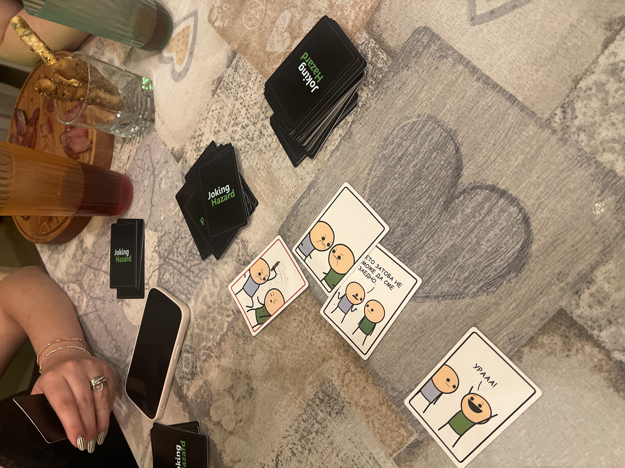 The image captures a casual indoor gathering, likely in a home in Plovdiv, Bulgaria, centered around a game of "Joking Hazard." A patterned tablecloth covers the table, upon which various items are arranged.
In the foreground, a person's left arm and hand are visible, holding game cards. The hand wears several bracelets and rings, and the fingernails are painted with a striped design. A white-cased smartphone rests nearby. Several stacks of "Joking Hazard" cards, identifiable by their black backs with green and white text, are spread across the table. Some cards are face up, revealing characteristic cartoon illustrations in the style of Cyanide & Happiness. One card shows three characters, with text that reads "ЕТО ЗАТОВА НЕ МОЖЕ ДА СМЕ ЗАЕДНО." (Bulgarian for "THAT'S WHY WE CAN'T BE TOGETHER."). Another card depicts two characters, with one exclaiming "УРААА!" (Bulgarian for "HOORAY!").
Snacks and drinks are also present. A clear glass holds several breadsticks or pretzels, positioned on a wooden cutting board that also contains slices of what appears to be cured meat or salami. Two ribbed cups, one green and one amber, filled with beverages, are also on the table. The overall scene suggests a relaxed social occasion, likely in the evening, focused on playing the card game and enjoying refreshments.