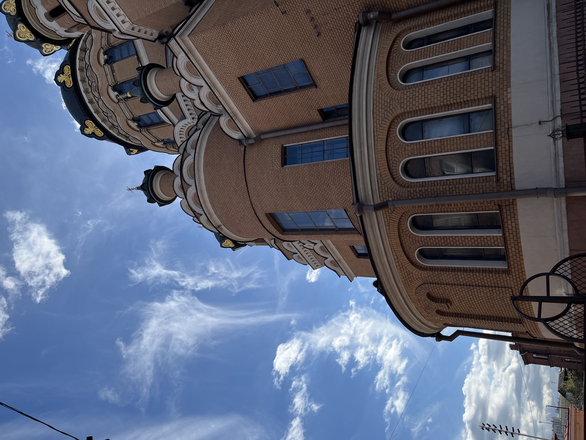 The image depicts a low-angle view of a building featuring a distinctive architectural style with brickwork and rounded elements. The building is constructed primarily of red brick and includes features such as arched windows. The top of the building incorporates dome-like structures that are decorated with gold-colored ornamentation.
Above the building is a bright blue sky populated with wispy white clouds. The image orientation has been rotated, giving the impression that the building is tilted.
