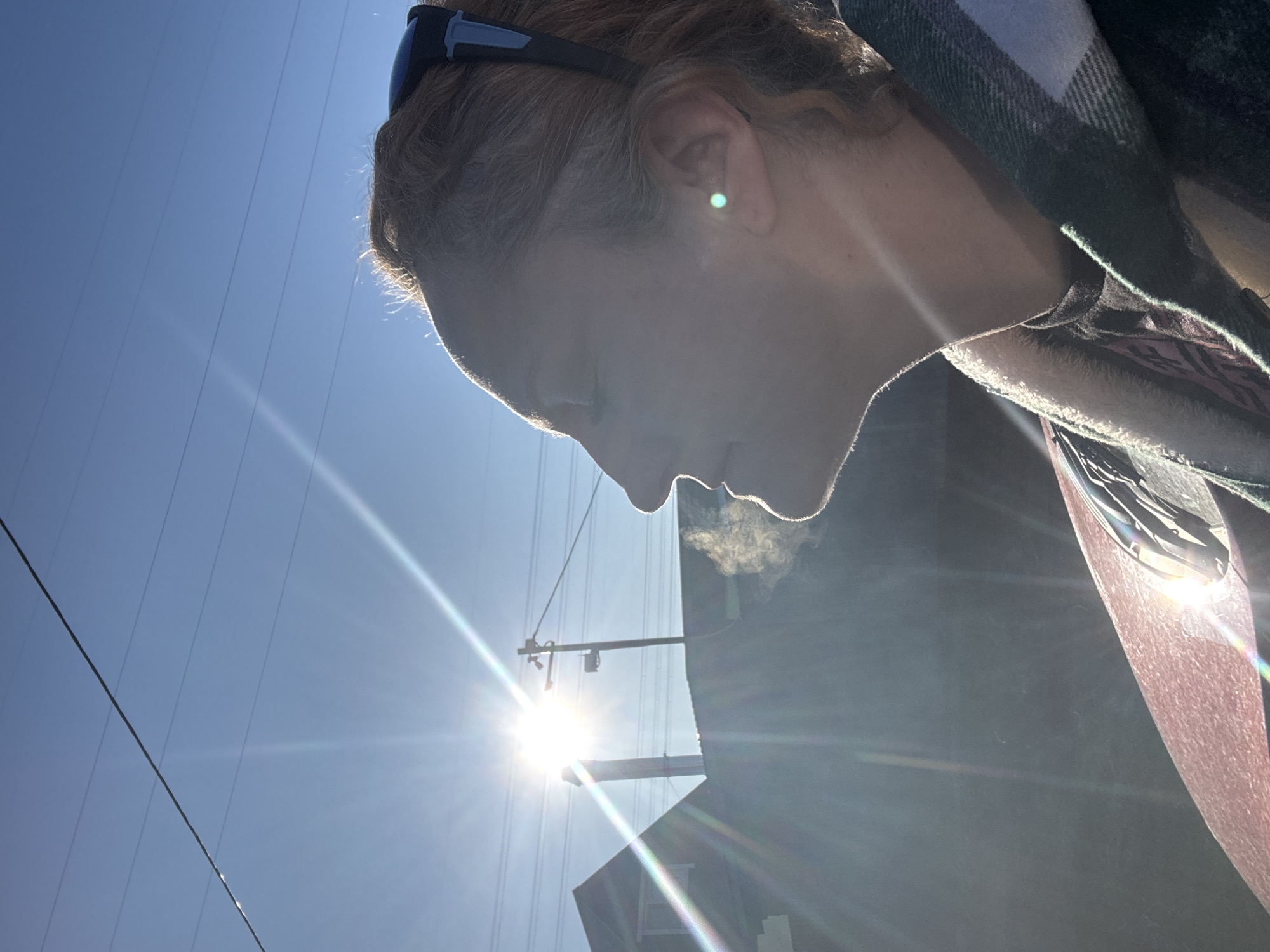 The image presents a backlit profile of a person outdoors, shot from a low angle. The subject's head, neck, and shoulder are visible on the right side of the frame, facing towards the lower left. The person has reddish-brown hair, styled back, and a pair of dark sunglasses with blue accents rest on their head. A small, bright green earring is visible on their ear. They are wearing a plaid garment, likely a scarf or jacket, with dark green/blue and white patterns.
A distinct cloud of white vapor or smoke is seen exhaling from the person's mouth, suggesting either cold weather causing visible breath or the act of vaping. A very bright, low sun is positioned in the upper right quadrant of the image, creating a strong lens flare that streaks across the clear blue sky and extends towards the person's body. The intense sunlight backlights the person, casting their face largely in shadow and highlighting the outline of their hair.
In the background, against the clear blue sky, several power lines run horizontally, intersecting with a utility pole. The dark silhouette of a building is partially visible on the far right. The overall scene suggests a bright but potentially cold daytime environment in an urban or suburban setting. There is no visible text in the image.